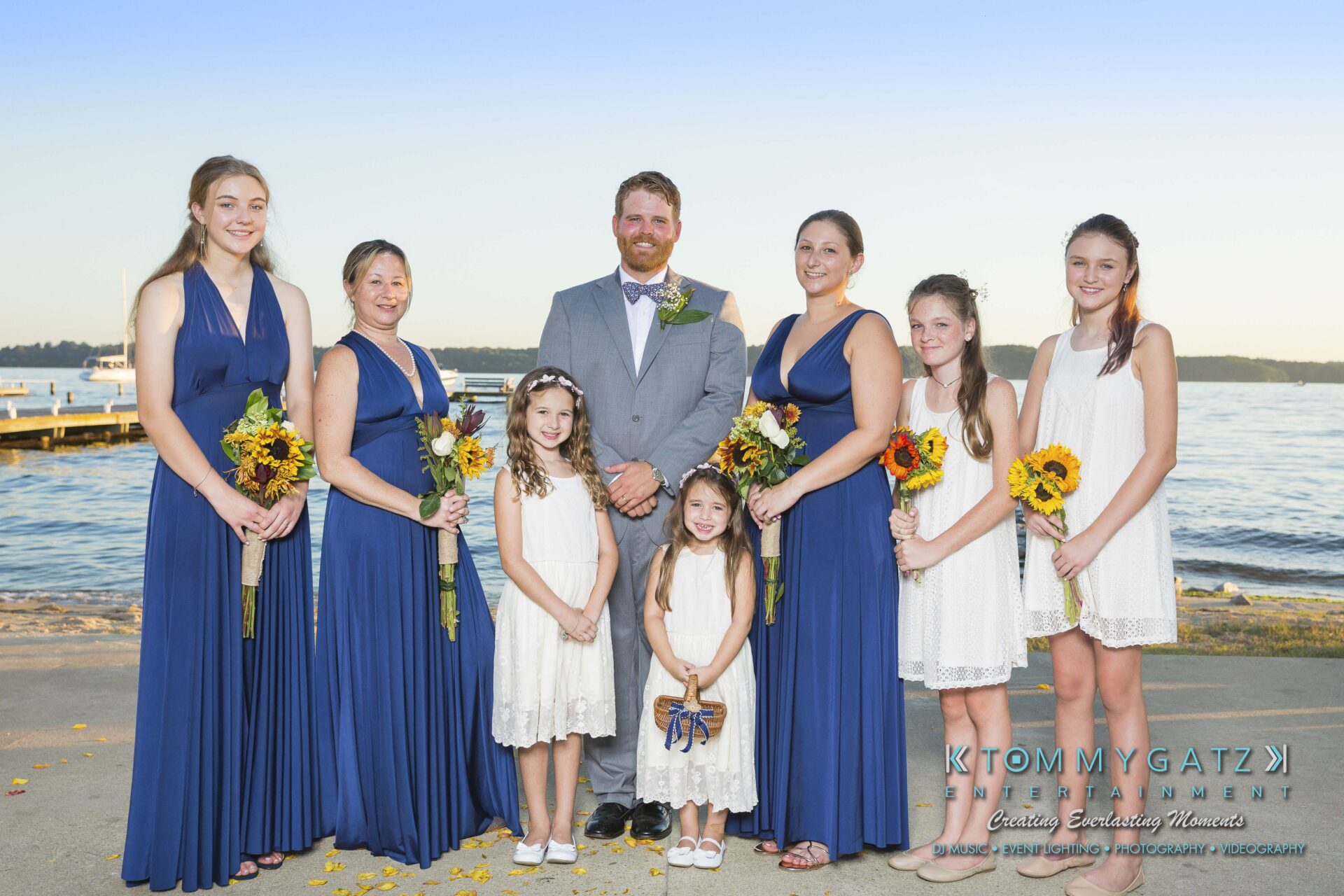 Ann-Marie-Alex-92317-FORMALS-Low-Res-WM-30 - TOMMY GATZ ENTERTAINMENT A group photo of a wedding party by a lakeside. Three women in blue dresses, three girls in white dresses, and a man in a suit stand smiling, holding sunflowers.