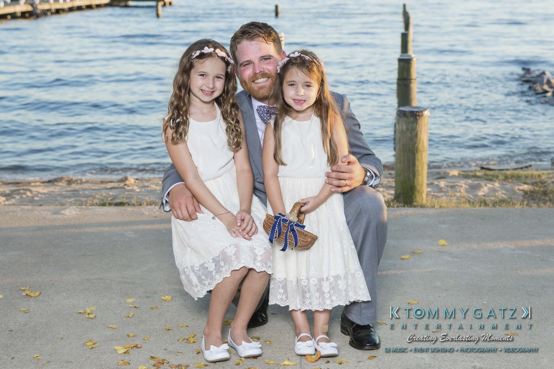 Ann-Marie-Alex-92317-FORMALS-Low-Res-WM-34 - TOMMY GATZ ENTERTAINMENT A man in a suit kneels by two young girls in white dresses, posing together by the water.