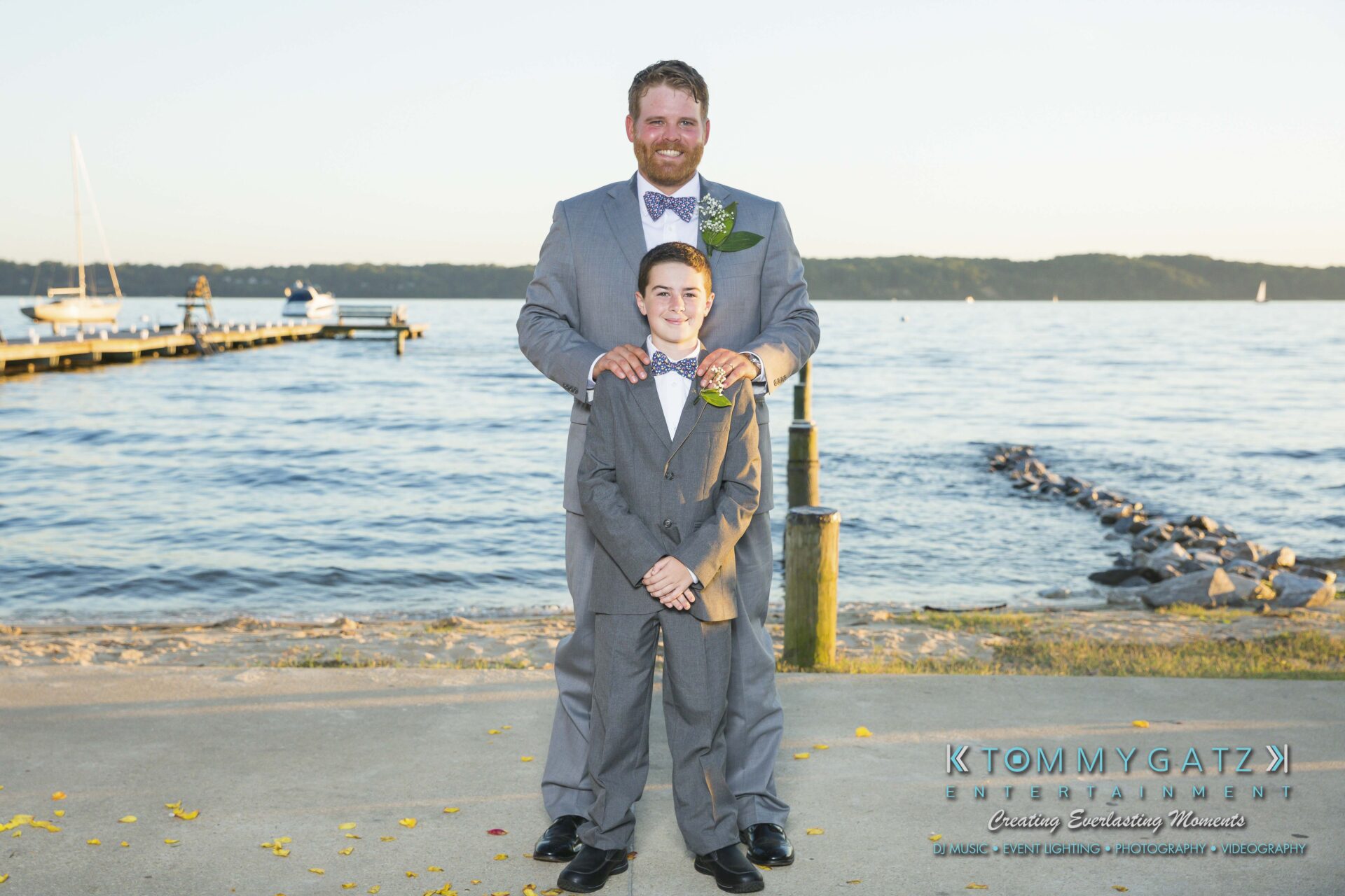 Ann-Marie-Alex-92317-FORMALS-Low-Res-WM-37 - TOMMY GATZ ENTERTAINMENT A man in a gray suit and a boy in a matching suit stand together by a calm waterfront. They are both smiling, with boats visible in the background.
