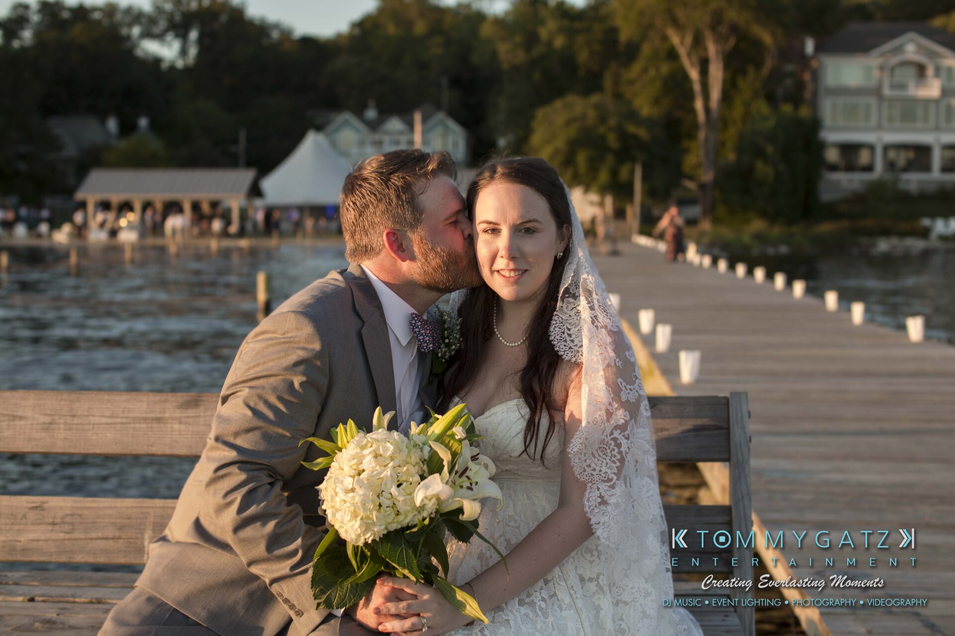 Ann-Marie-Alex-92317-FORMALS-Low-Res-WM-39 - TOMMY GATZ ENTERTAINMENT A groom kisses a bride's cheek as they sit on a bench by a lake, with a wooden dock and houses in the background. The bride holds a bouquet of white flowers.