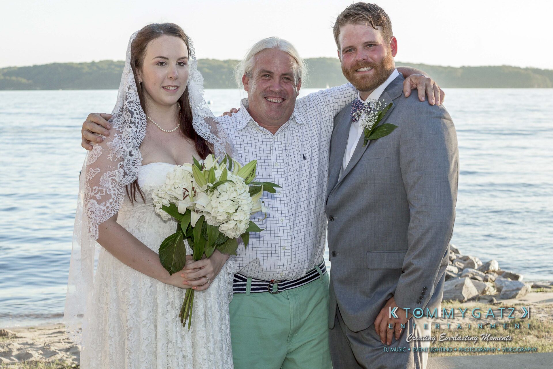 Ann-Marie-Alex-92317-FORMALS-Low-Res-WM-4 - TOMMY GATZ ENTERTAINMENT A bride, a groom, and an older man pose together by a body of water. The bride holds a bouquet and wears a lace veil.