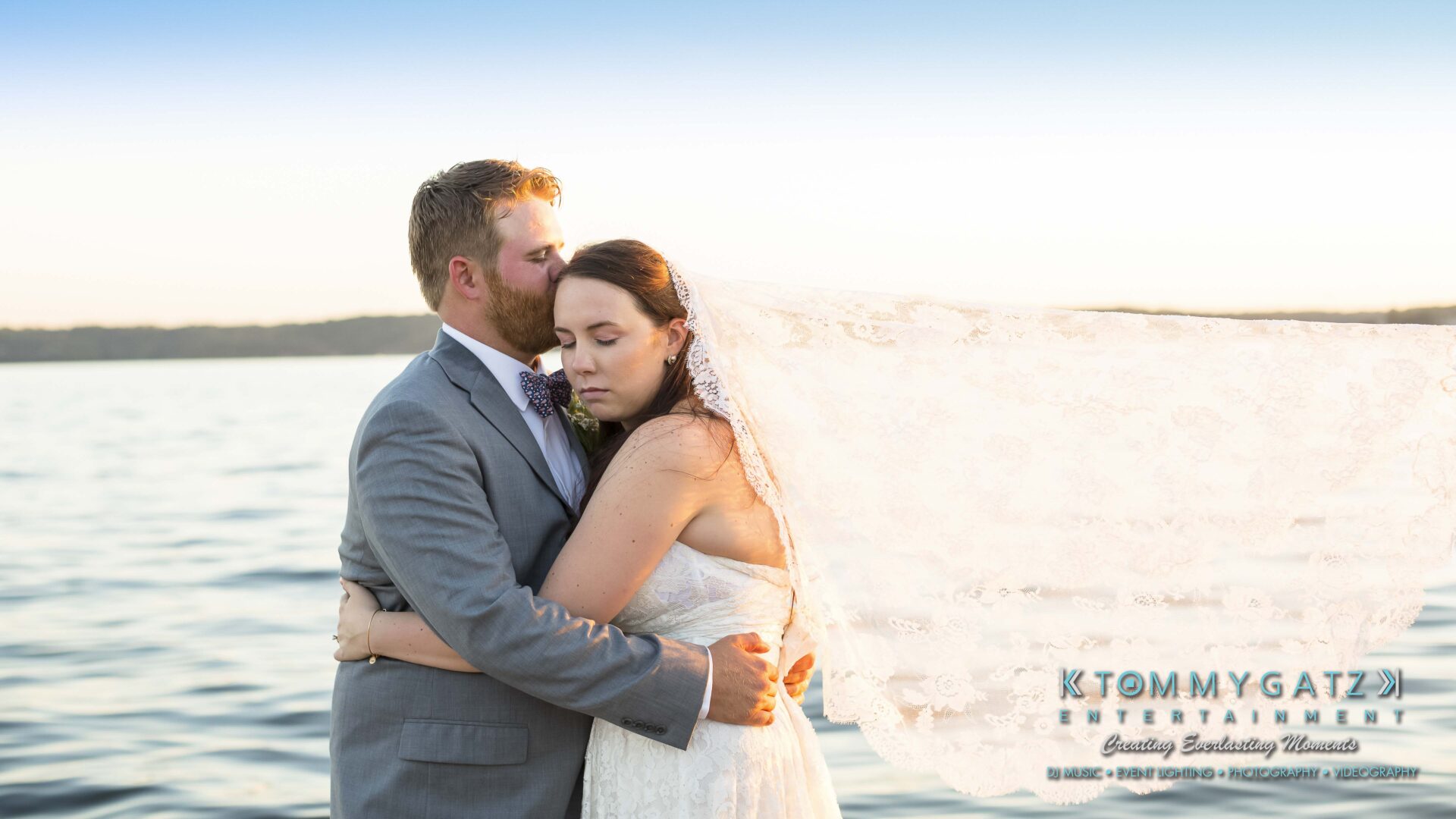 Ann-Marie-Alex-92317-FORMALS-Low-Res-WM-47 - TOMMY GATZ ENTERTAINMENT A bride and groom embrace on a lake shore at sunset, with the bride's veil flowing in the breeze.