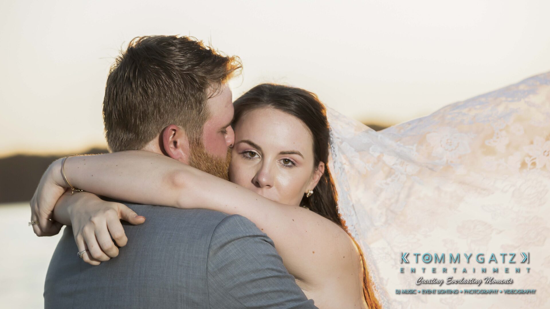 Ann-Marie-Alex-92317-FORMALS-Low-Res-WM-53 - TOMMY GATZ ENTERTAINMENT A couple embraces beside a body of water, the woman's arms wrapped around the man's shoulders, with a sunset in the background.