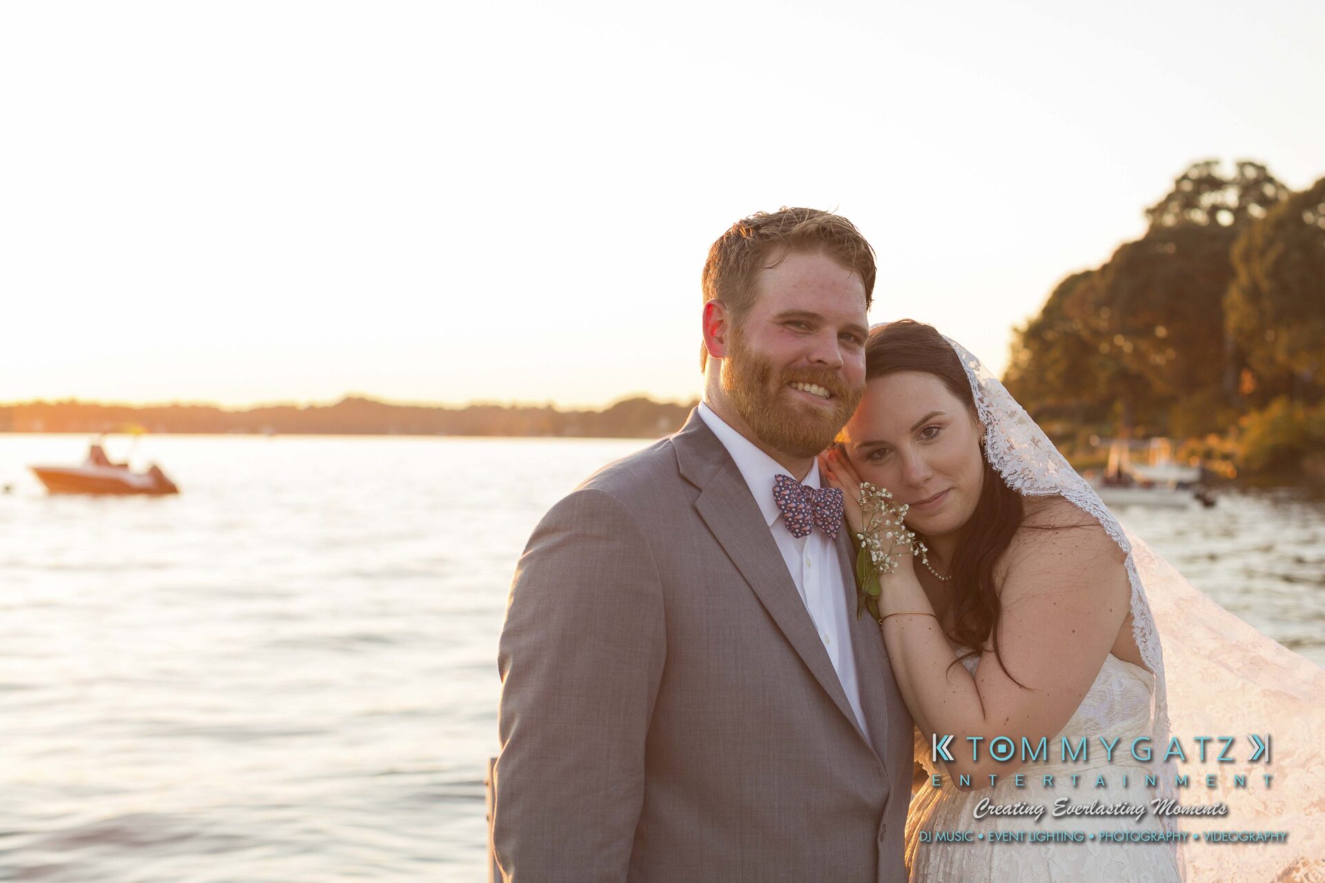 Ann-Marie-Alex-92317-FORMALS-Low-Res-WM-56 - TOMMY GATZ ENTERTAINMENT A couple poses by a lakeside at sunset. The groom is in a gray suit and the bride wears a veil and holds a bouquet. A boat is visible in the water.