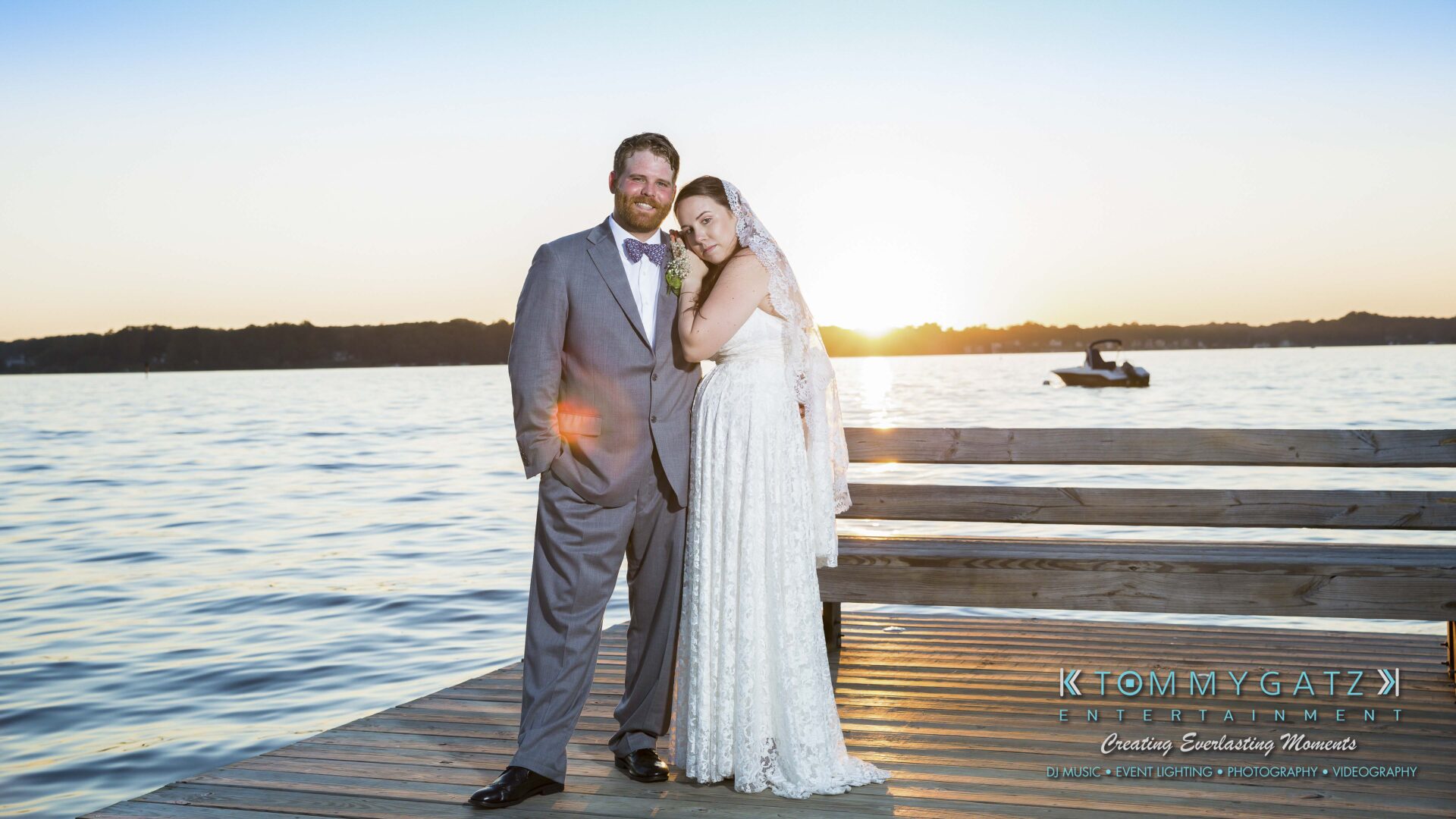 Ann-Marie-Alex-92317-FORMALS-Low-Res-WM-59 - TOMMY GATZ ENTERTAINMENT A bride and groom stand on a wooden dock by a lake at sunset. The bride leans on the groom, and a small boat is visible in the background.