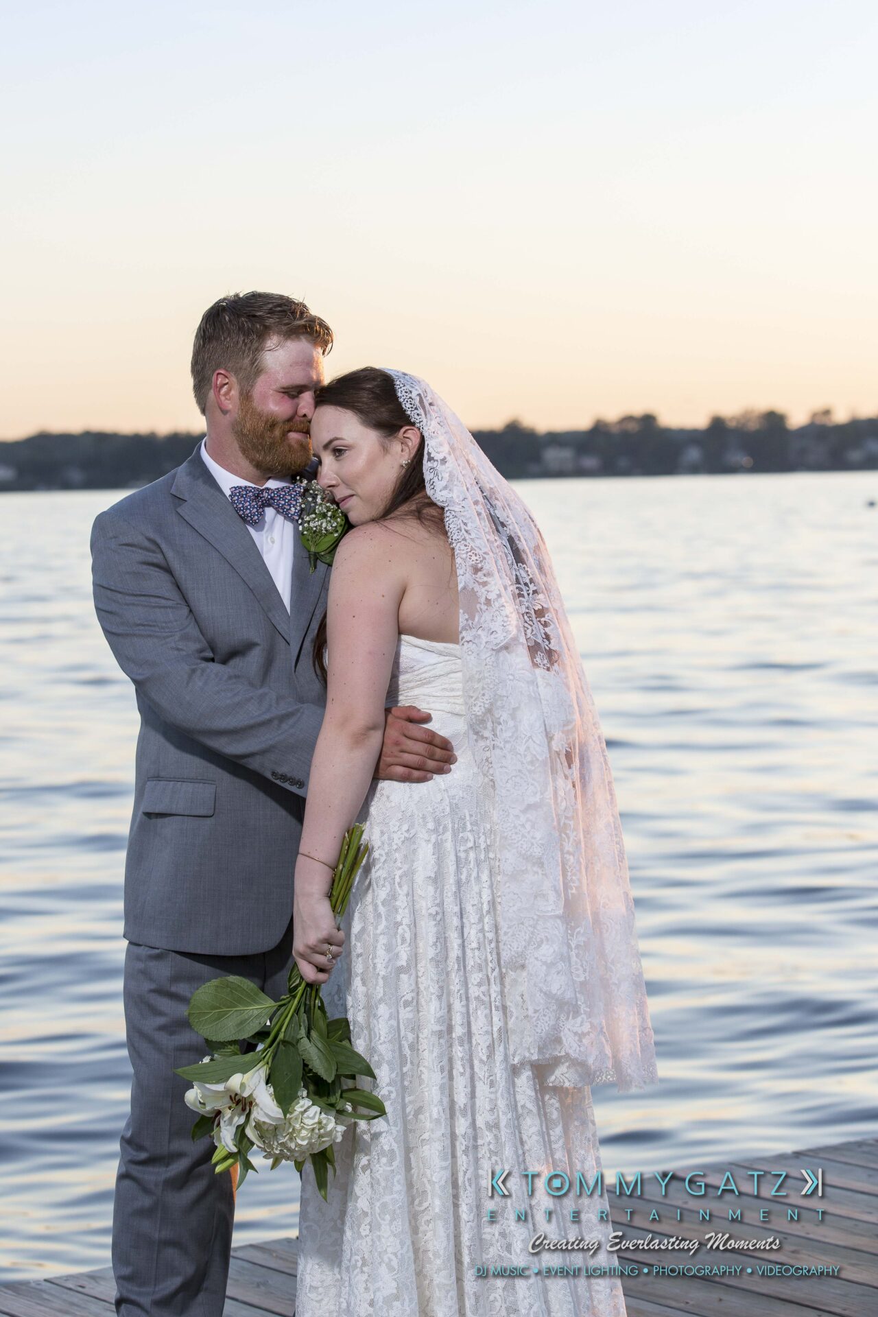 Ann-Marie-Alex-92317-FORMALS-Low-Res-WM-64 - TOMMY GATZ ENTERTAINMENT A bride and groom stand together on a dock by a lake, with the groom wearing a suit and the bride in a white dress holding flowers.