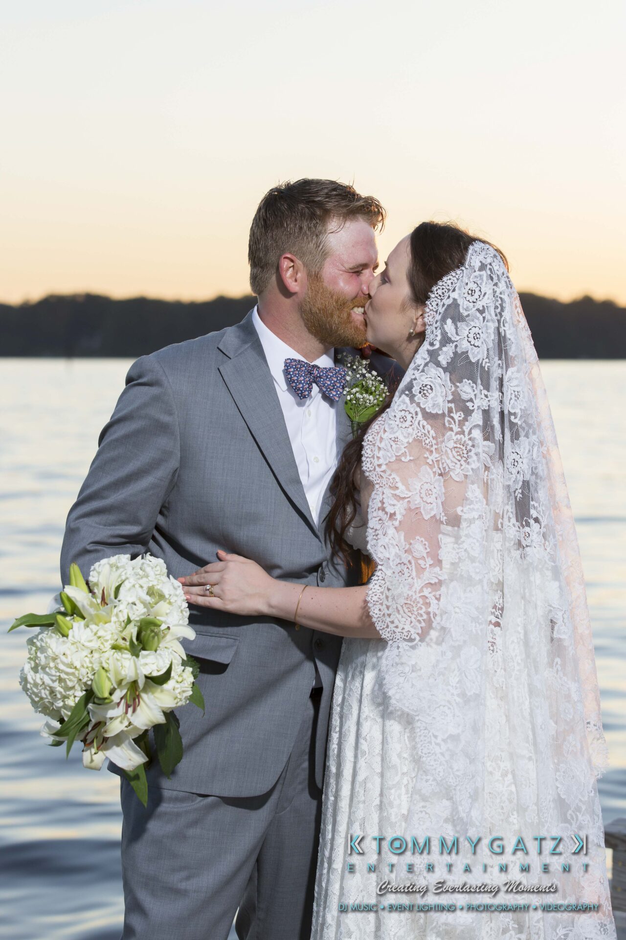 Ann-Marie-Alex-92317-FORMALS-Low-Res-WM-66 - TOMMY GATZ ENTERTAINMENT Bride and groom kissing by a lake at sunset, with the bride holding a bouquet of white flowers.