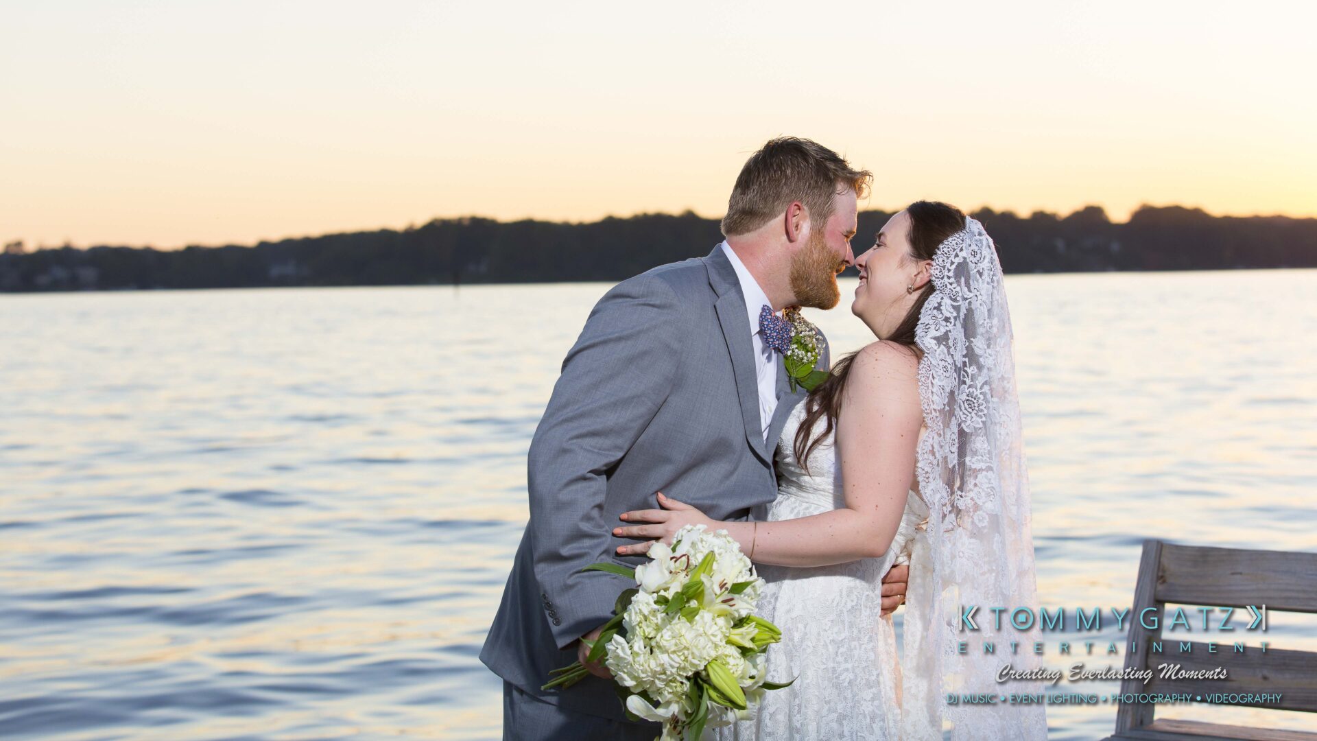 Ann-Marie-Alex-92317-FORMALS-Low-Res-WM-67 - TOMMY GATZ ENTERTAINMENT A bride and groom stand by a lake at sunset, with the groom holding a bouquet.