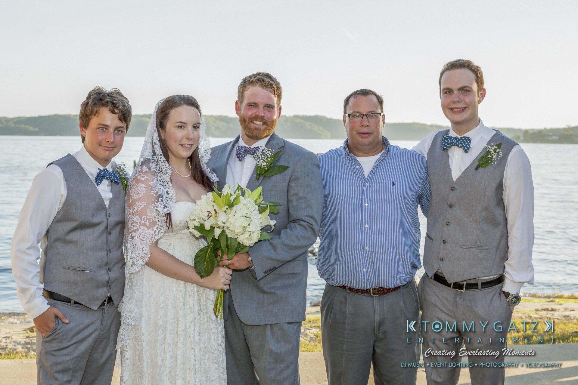 Ann-Marie-Alex-92317-FORMALS-Low-Res-WM-68 - TOMMY GATZ ENTERTAINMENT A wedding party stands outdoors by a waterfront. Three men wear grey suits, two with bow ties. The bride holds a bouquet, and another man wears a blue shirt and trousers.