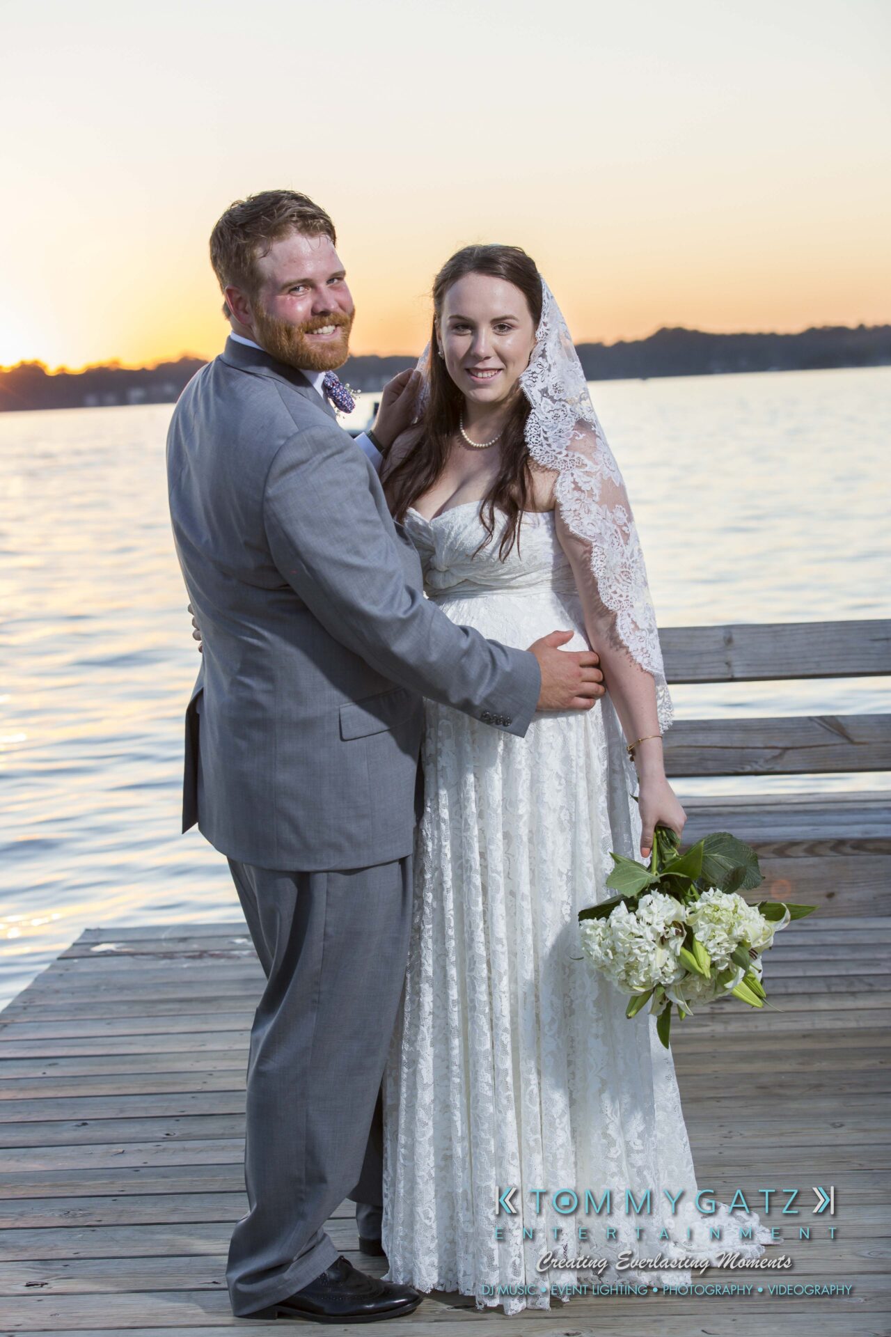 Ann-Marie-Alex-92317-FORMALS-Low-Res-WM-73 - TOMMY GATZ ENTERTAINMENT Bride and groom in wedding attire stand on a wooden dock by the water, smiling and embracing.