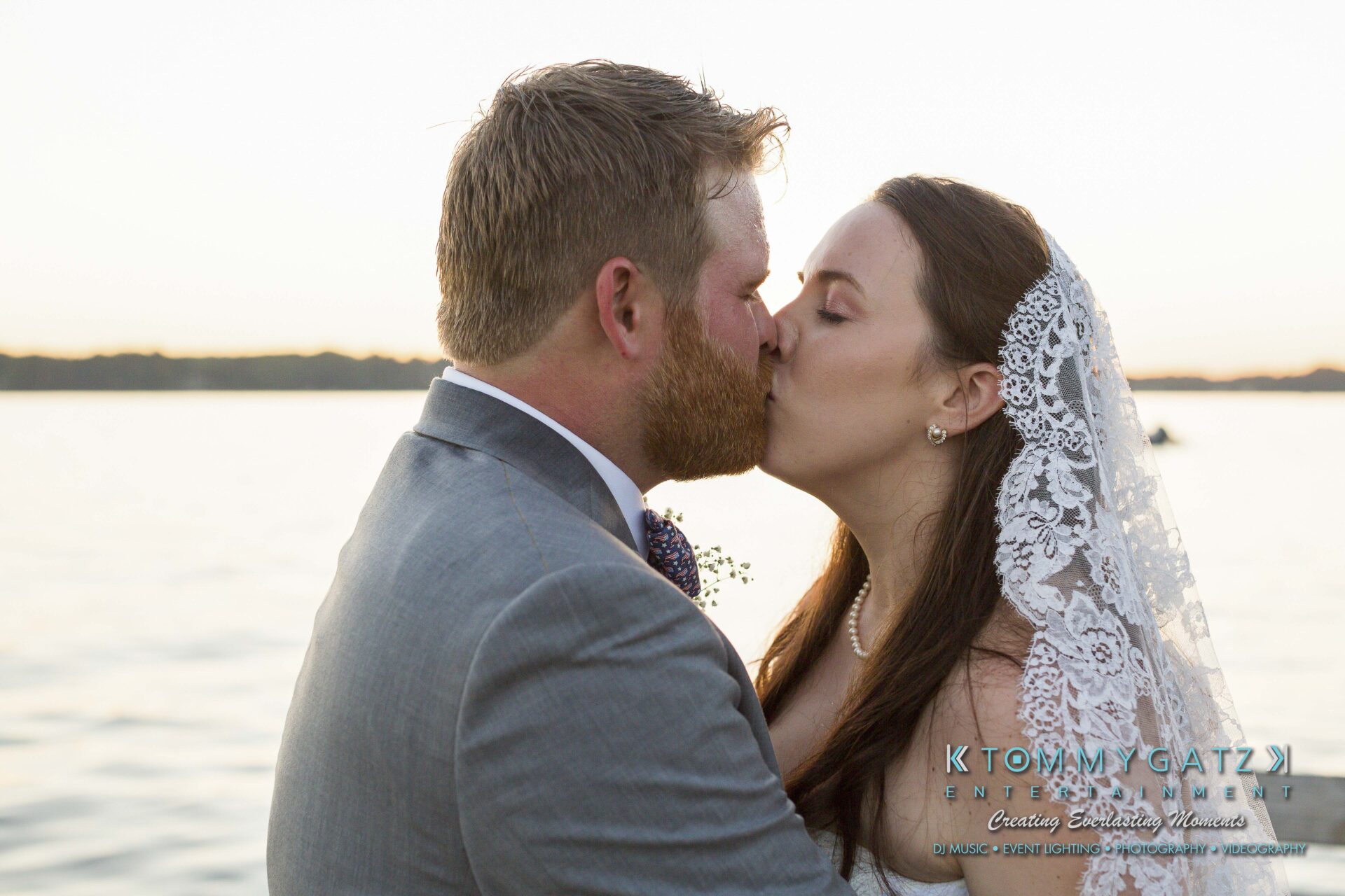 Ann-Marie-Alex-92317-FORMALS-Low-Res-WM-74 - TOMMY GATZ ENTERTAINMENT A bride and groom kiss by a lake at sunset. The bride wears a lace veil and pearl necklace, and the groom is in a gray suit. The lake and horizon are visible in the background.