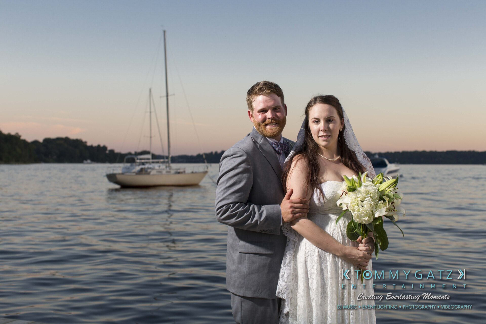 Ann-Marie-Alex-92317-FORMALS-Low-Res-WM-76 - TOMMY GATZ ENTERTAINMENT A bride and groom stand by a lake with a sailboat in the background, under a twilight sky. The bride holds a bouquet of flowers.