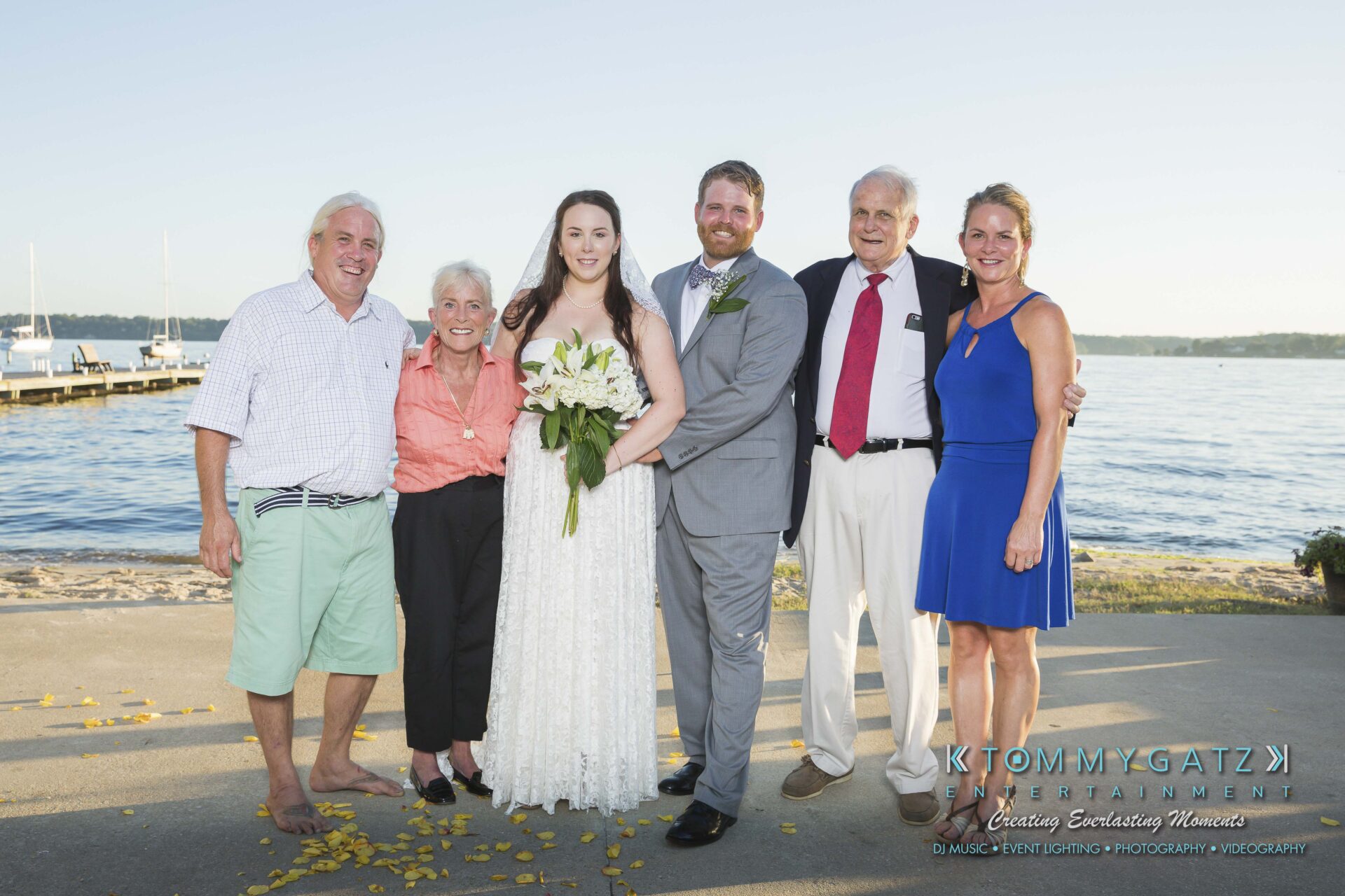 Ann-Marie-Alex-92317-FORMALS-Low-Res-WM-8 - TOMMY GATZ ENTERTAINMENT A bride and groom stand with four people by a waterfront, with a dock and boats visible in the background.