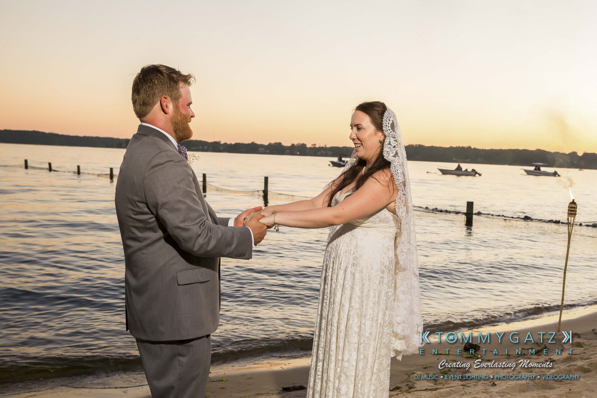 Ann-Marie-Alex-92317-FORMALS-Low-Res-WM-80 - TOMMY GATZ ENTERTAINMENT A bride and groom hold hands standing on a sandy beach, with a body of water and several boats in the background at sunset.