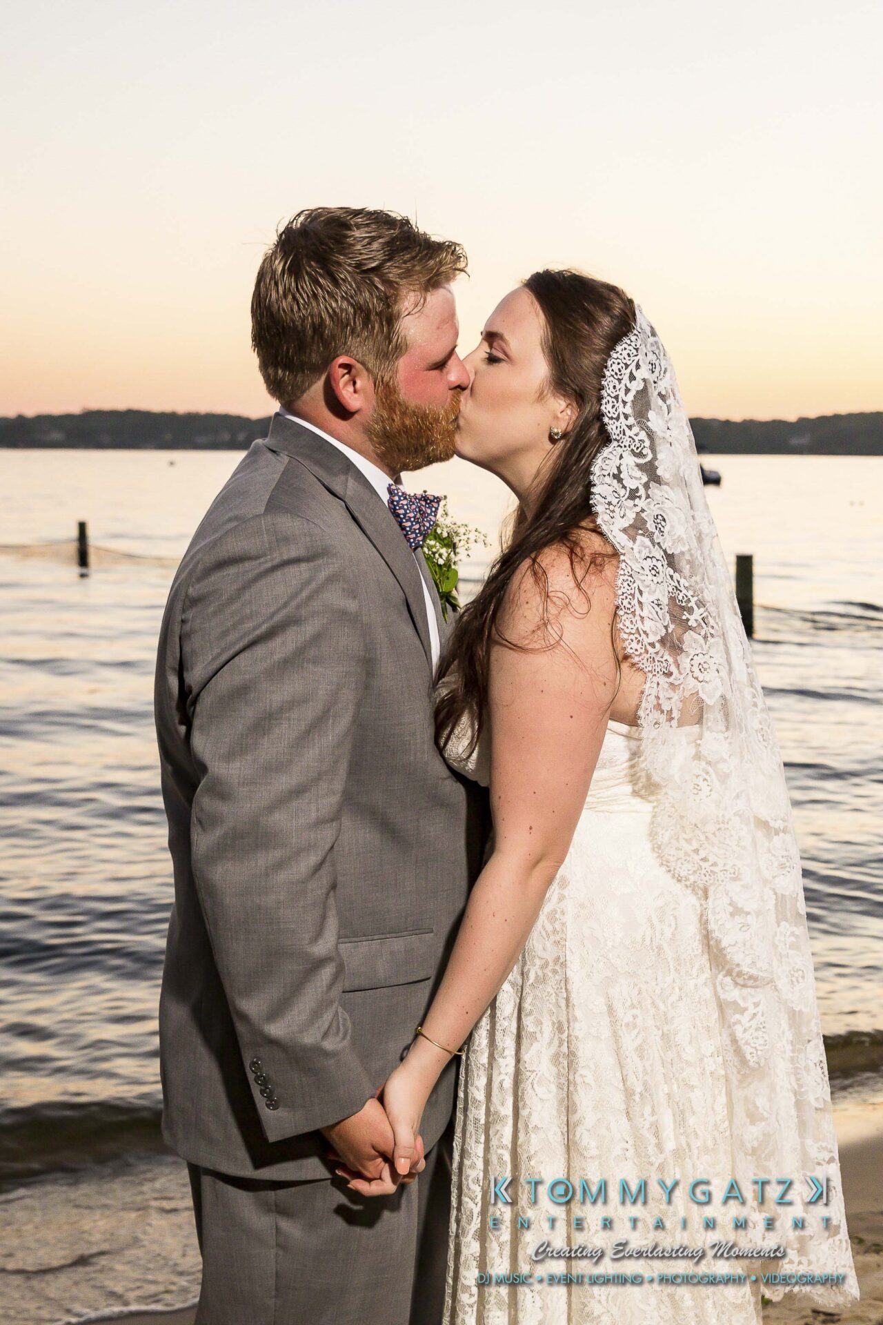 Ann-Marie-Alex-92317-FORMALS-Low-Res-WM-82 - TOMMY GATZ ENTERTAINMENT A bride and groom share a kiss by a lakeside at sunset, holding hands.
