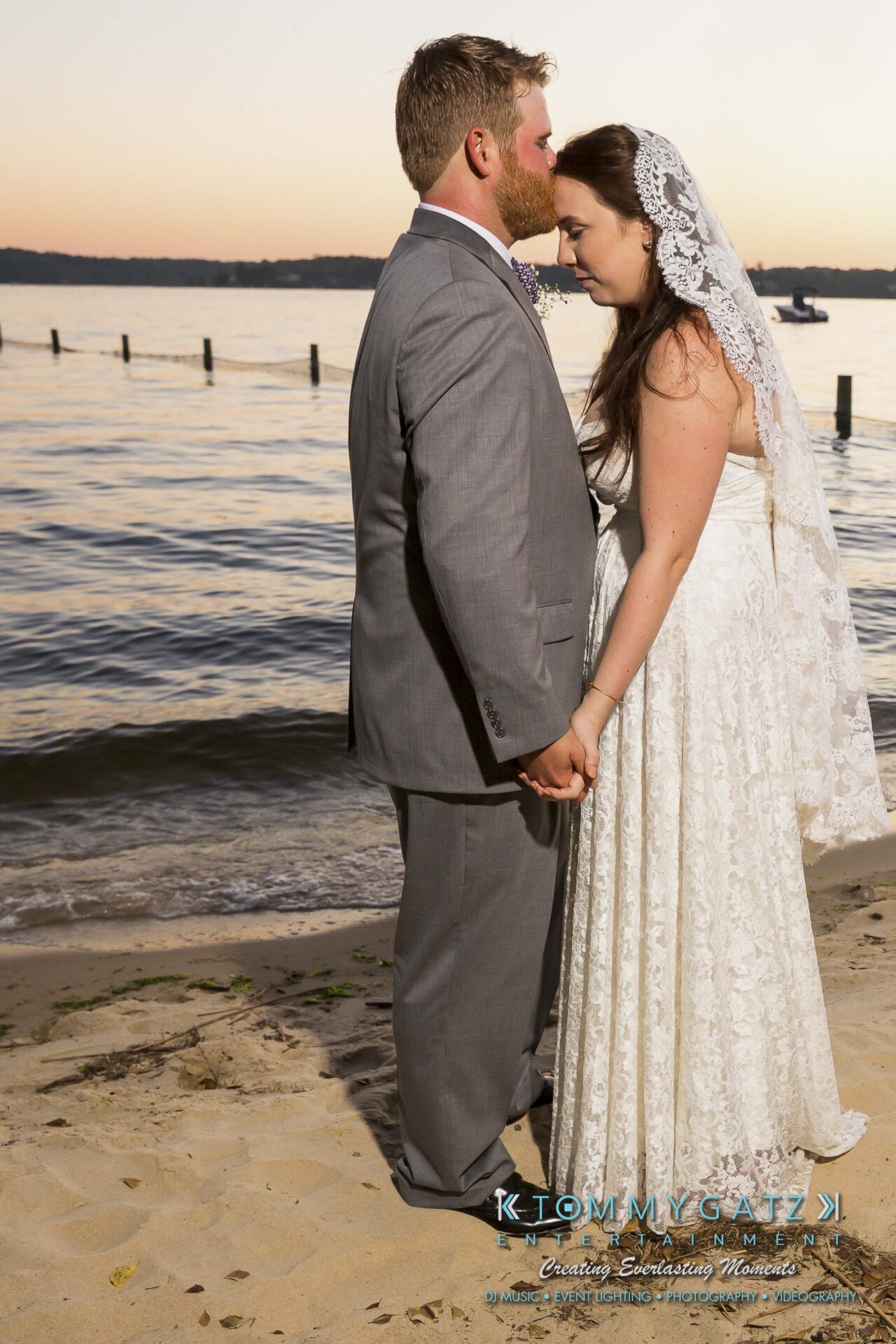 Ann-Marie-Alex-92317-FORMALS-Low-Res-WM-83 - TOMMY GATZ ENTERTAINMENT A bride and groom stand by the water at sunset, holding hands and facing each other. The bride wears a lace gown and veil, and the groom is in a gray suit. Silent serenity surrounds them.