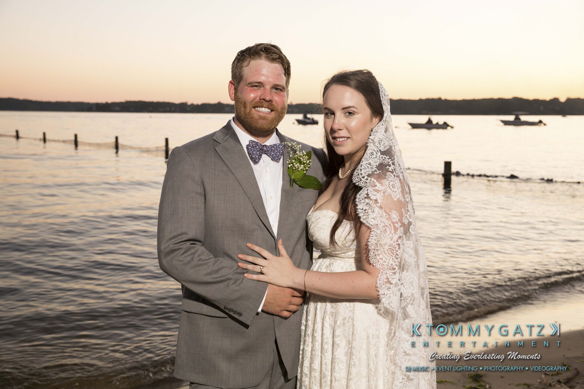 Ann-Marie-Alex-92317-FORMALS-Low-Res-WM-84 - TOMMY GATZ ENTERTAINMENT A bride and groom in formal attire pose by a lake at sunset. The bride wears a lace veil, and the groom wears a gray suit with a bow tie.