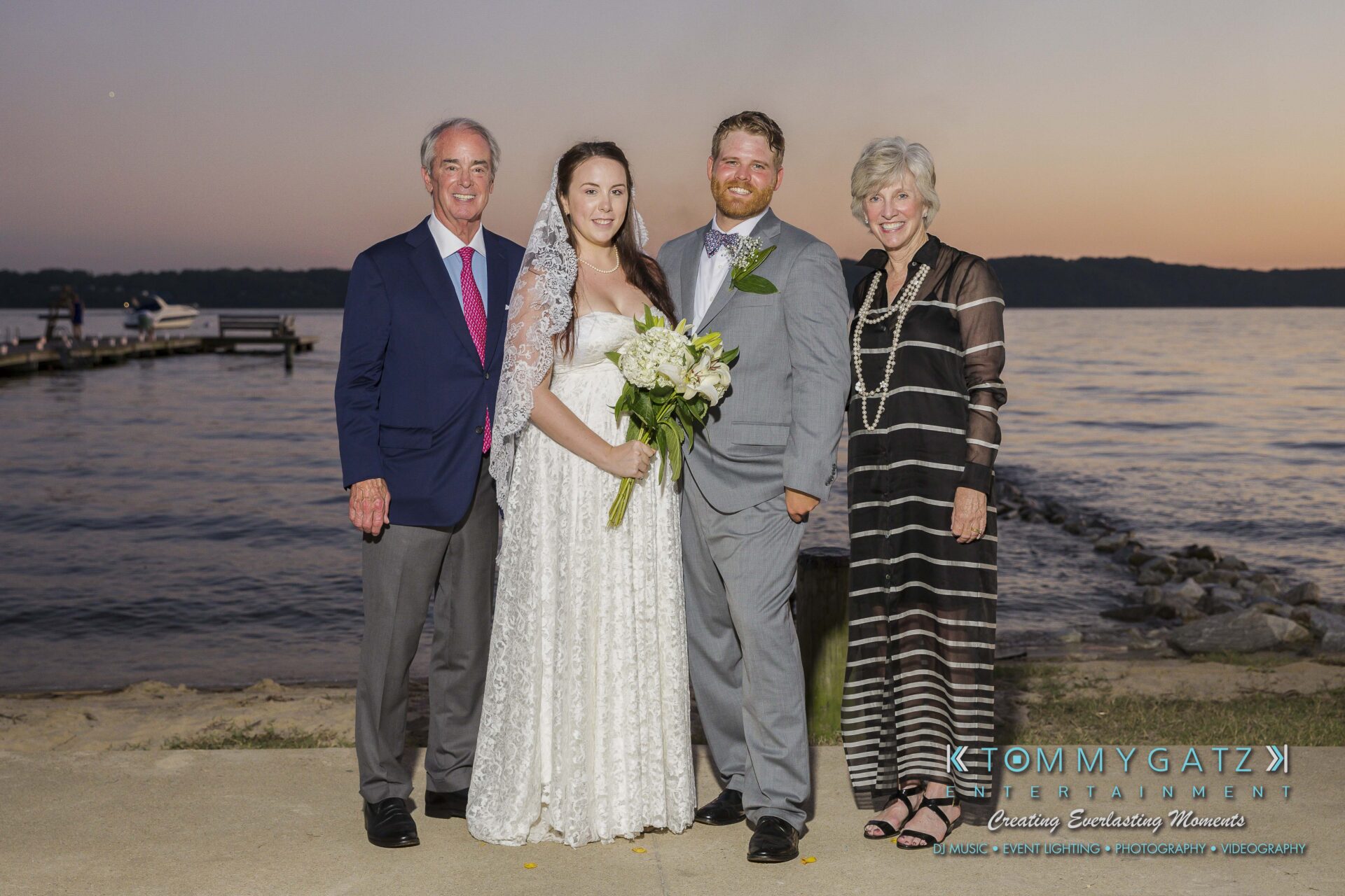 Ann-Marie-Alex-92317-FORMALS-Low-Res-WM-89 - TOMMY GATZ ENTERTAINMENT A bride and groom stand with two older adults near a lakeside at sunset, dressed in formal attire.