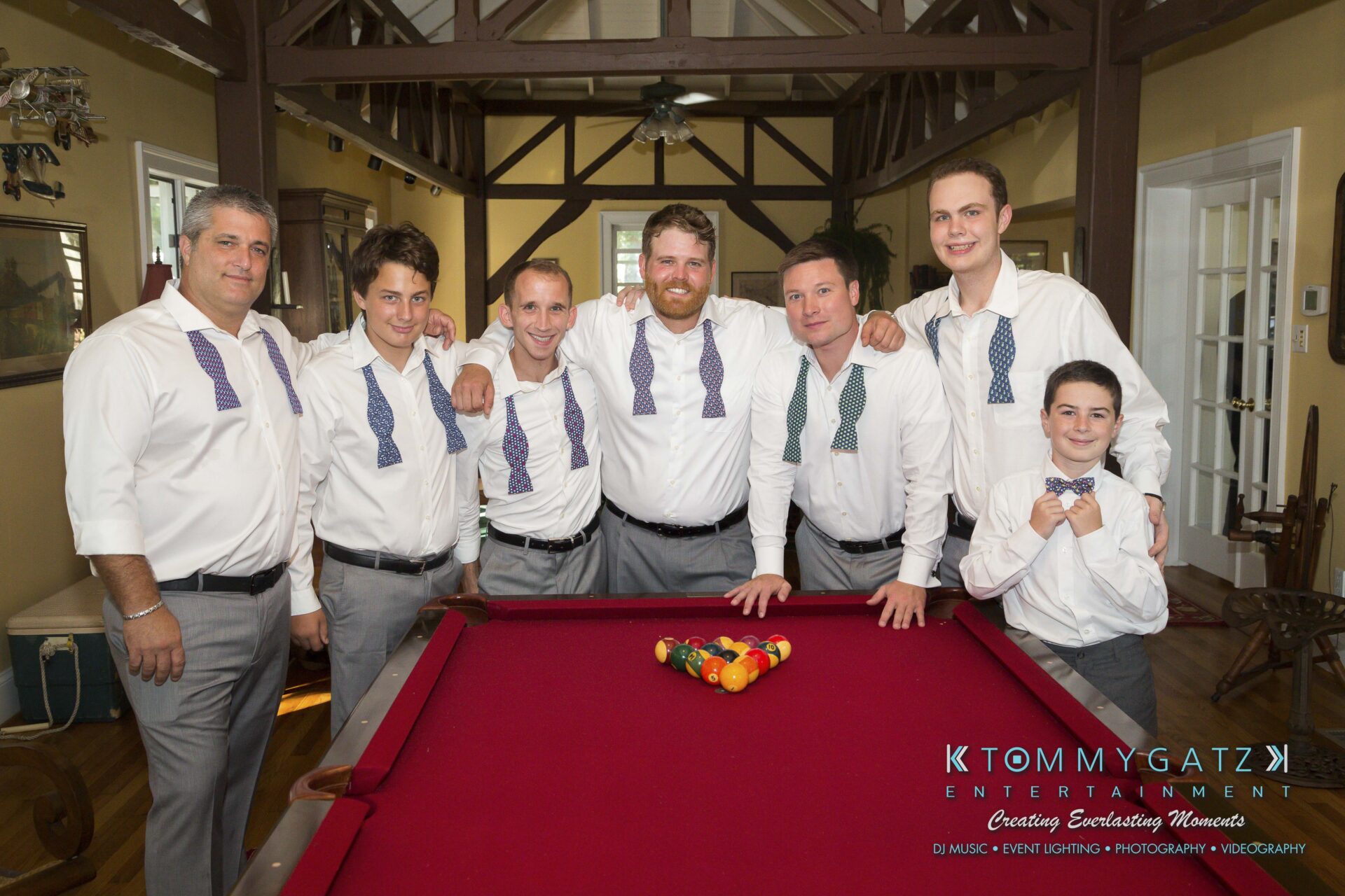 Ann-Marie-Alex-92317-PREP-Low-Res-WM-107 - TOMMY GATZ ENTERTAINMENT Seven people posing around a red pool table, wearing white shirts and bow ties, in a room with wooden beams.