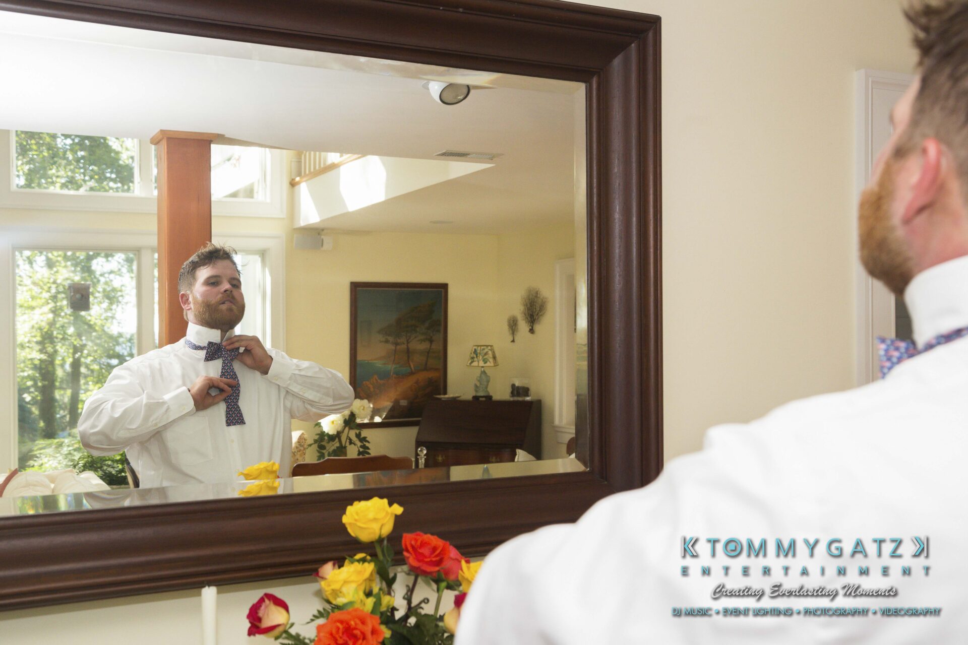 Ann-Marie-Alex-92317-PREP-Low-Res-WM-123 - TOMMY GATZ ENTERTAINMENT A man adjusts his bow tie in front of a mirror in a brightly lit room with flowers on a table.
