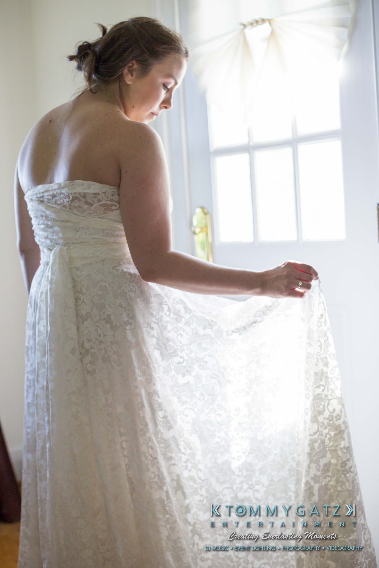Ann-Marie-Alex-92317-PREP-Low-Res-WM-131 - TOMMY GATZ ENTERTAINMENT A woman in a lace wedding dress stands by a sunlit door, holding the dress's fabric and looking down.