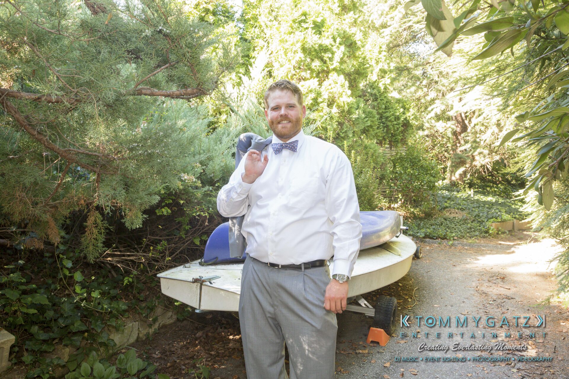 Ann-Marie-Alex-92317-PREP-Low-Res-WM-135 - TOMMY GATZ ENTERTAINMENT A man in a white shirt and bow tie stands outdoors, smiling, with a canoe and trees in the background.