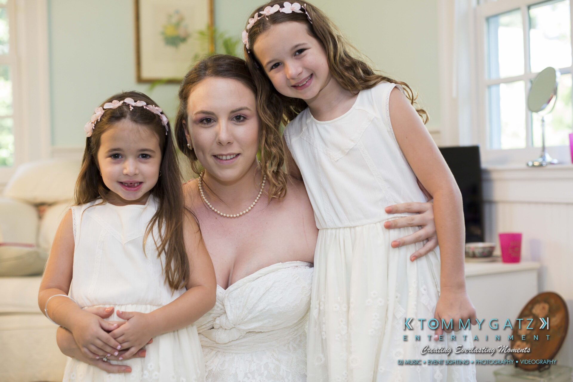 Ann-Marie-Alex-92317-PREP-Low-Res-WM-142 - TOMMY GATZ ENTERTAINMENT A woman in a white dress poses with two young girls, also in white dresses, indoors. The room has pastel-colored decor and natural light.
