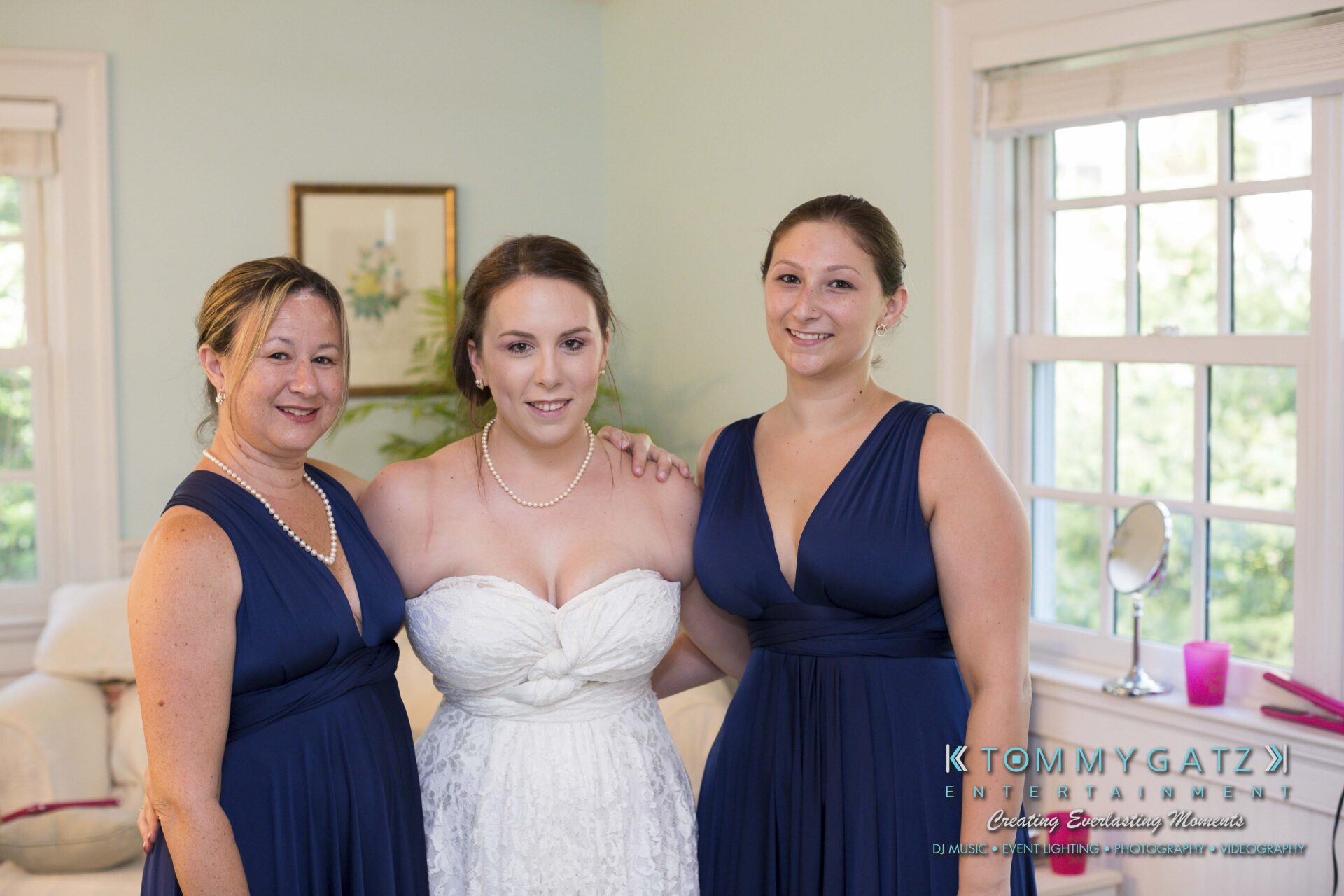Ann-Marie-Alex-92317-PREP-Low-Res-WM-145 - TOMMY GATZ ENTERTAINMENT Three women stand together in a room. The woman in the center is wearing a white dress, while the other two are in blue dresses. They are smiling and posing for the photo.
