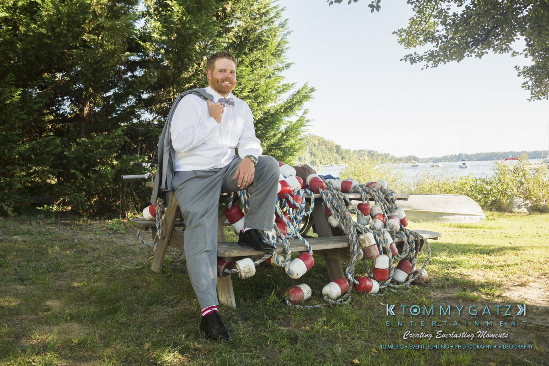 Ann-Marie-Alex-92317-PREP-Low-Res-WM-151 - TOMMY GATZ ENTERTAINMENT Man in a white shirt and gray pants sitting on a picnic table surrounded by nautical ropes and buoys, with trees and a lake in the background.