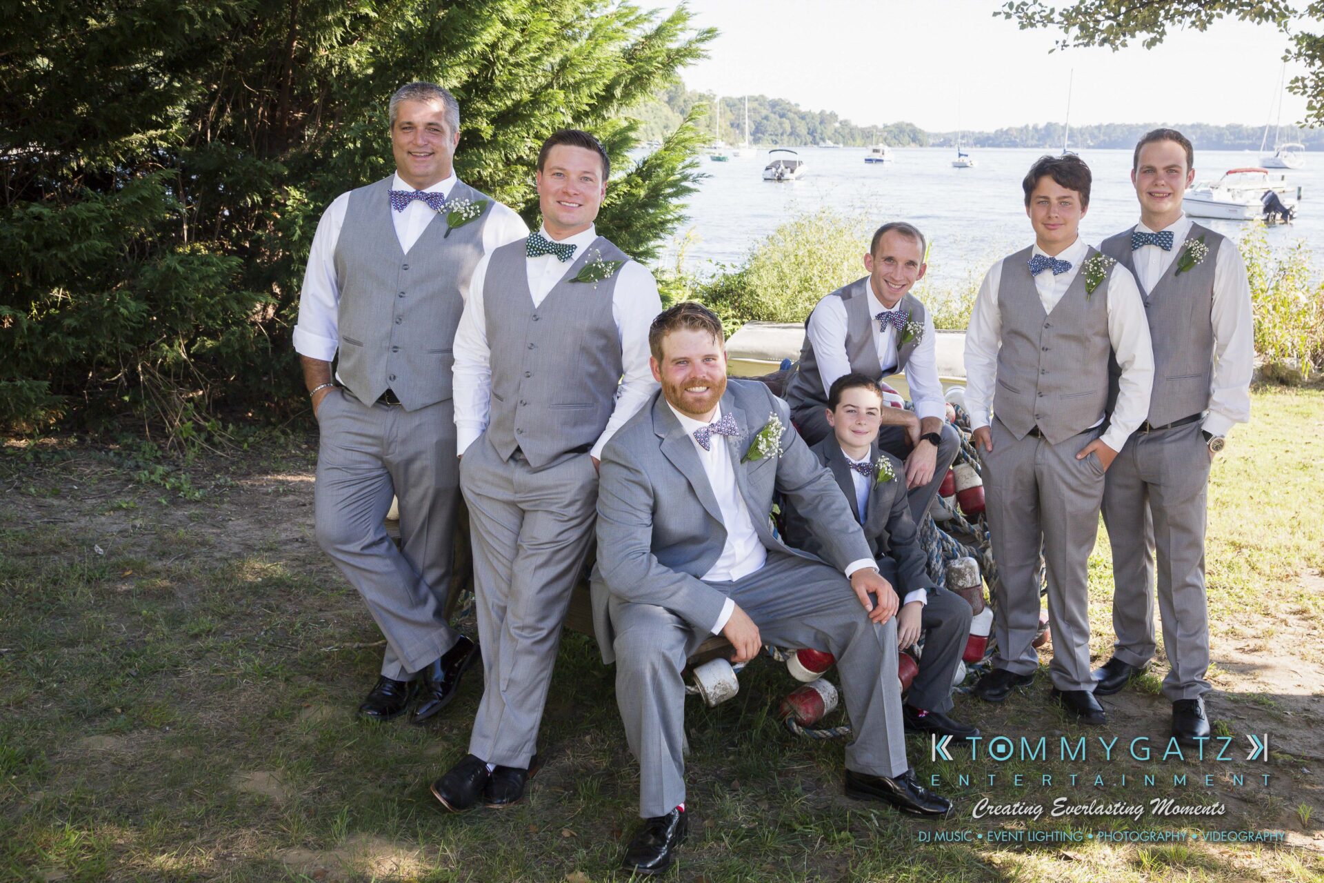 Ann-Marie-Alex-92317-PREP-Low-Res-WM-19 - TOMMY GATZ ENTERTAINMENT A group of men in gray suits with bow ties pose outdoors near a lake. Some are standing, and others are seated on a log, with trees and boats visible in the background.