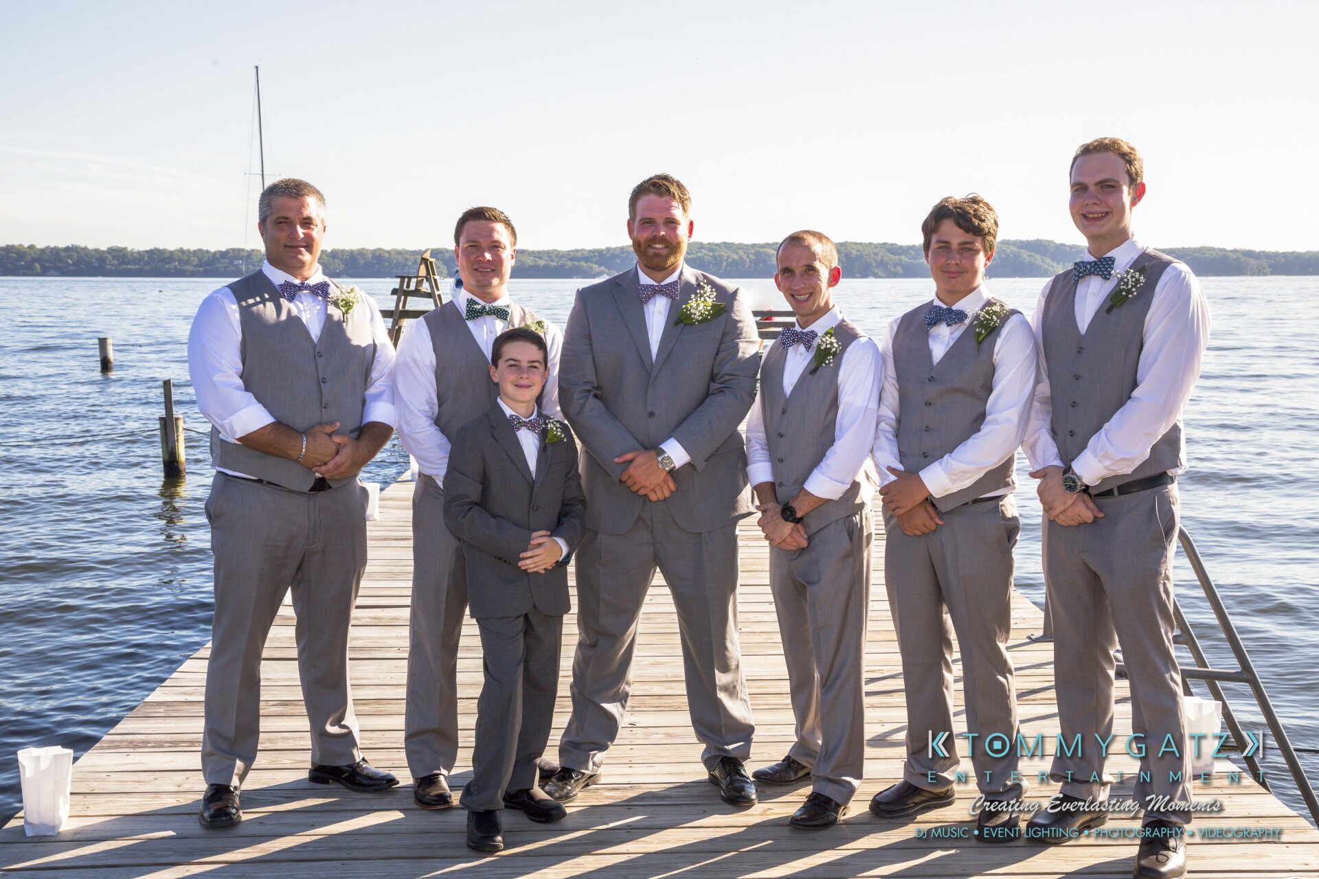 Ann-Marie-Alex-92317-PREP-Low-Res-WM-23 - TOMMY GATZ ENTERTAINMENT A group of seven men in suits, including a young boy, stand on a dock by a lake. They wear gray vests and bow ties, with the central figure in a full suit.