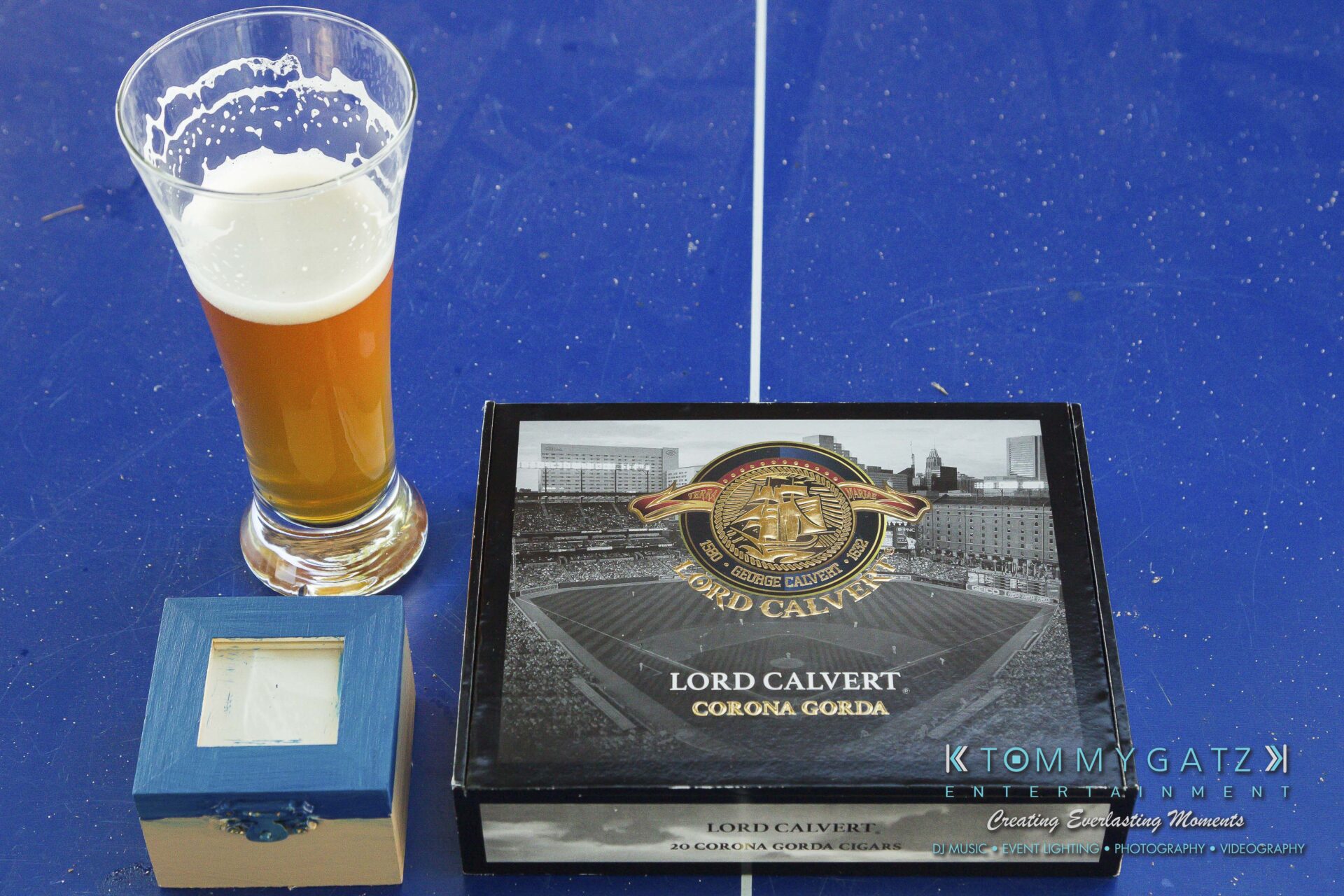Ann-Marie-Alex-92317-PREP-Low-Res-WM-5 - TOMMY GATZ ENTERTAINMENT A glass of beer, a small blue box, and a Lord Calvert cigar box on a blue surface with "KTOMMY GATZ ENTERTAINMENT" text.