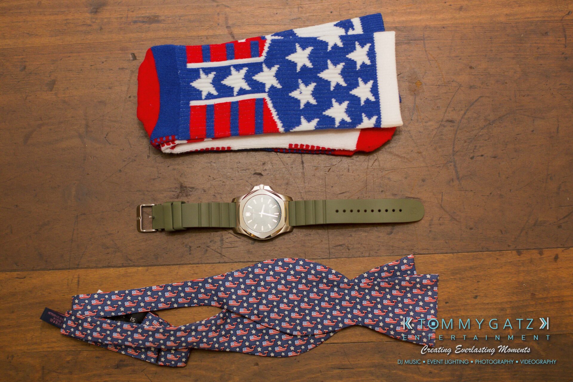 Ann-Marie-Alex-92317-PREP-Low-Res-WM-76 - TOMMY GATZ ENTERTAINMENT Patriotic socks, a green watch, and a bow tie with flag design on a wooden surface.