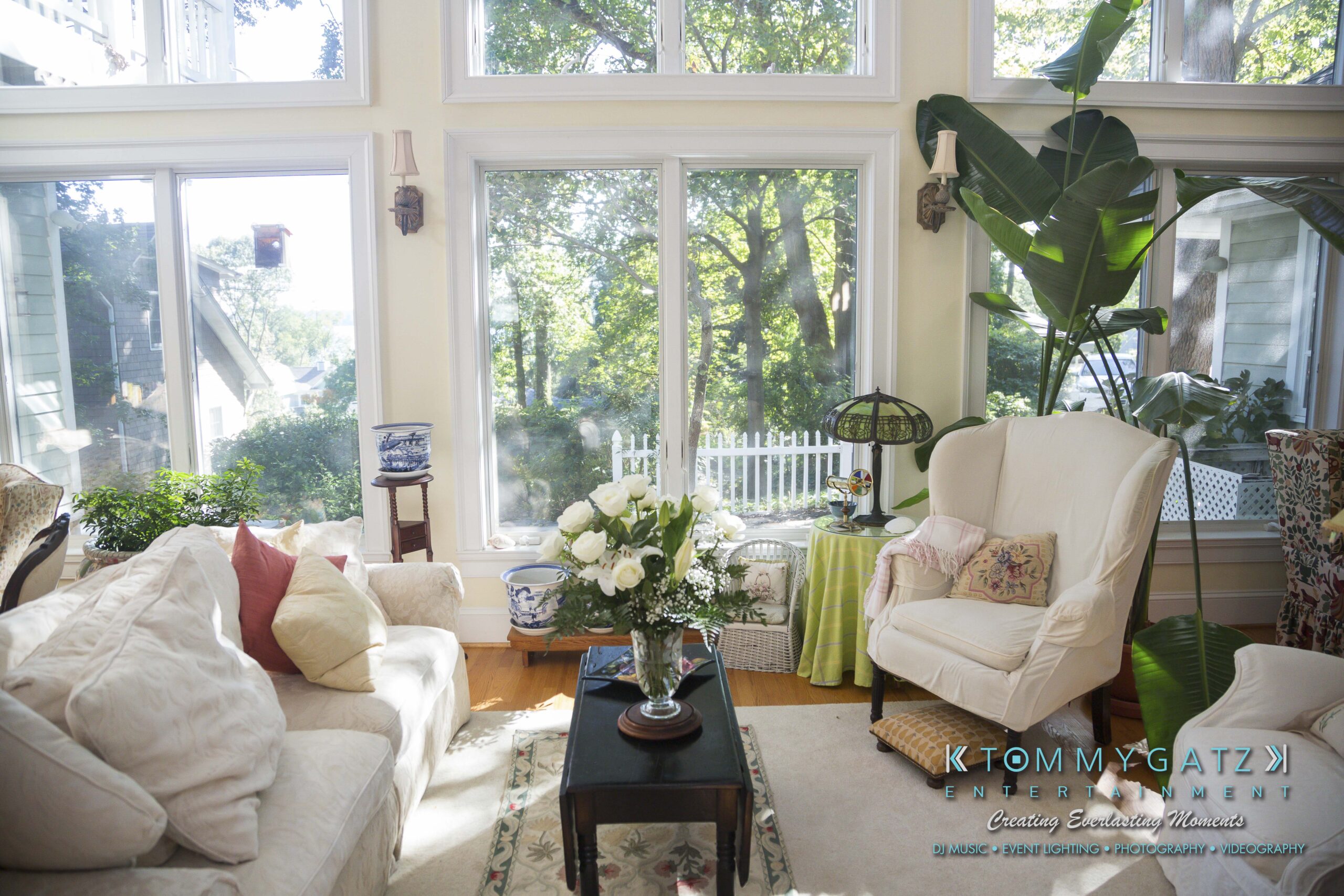 Ann-Marie-Alex-92317-PREP-Low-Res-WM-85 - TOMMY GATZ ENTERTAINMENT A sunlit living room with large windows, white armchairs, a sofa, and a wooden coffee table with a vase of flowers. Greenery outside is visible through the windows.