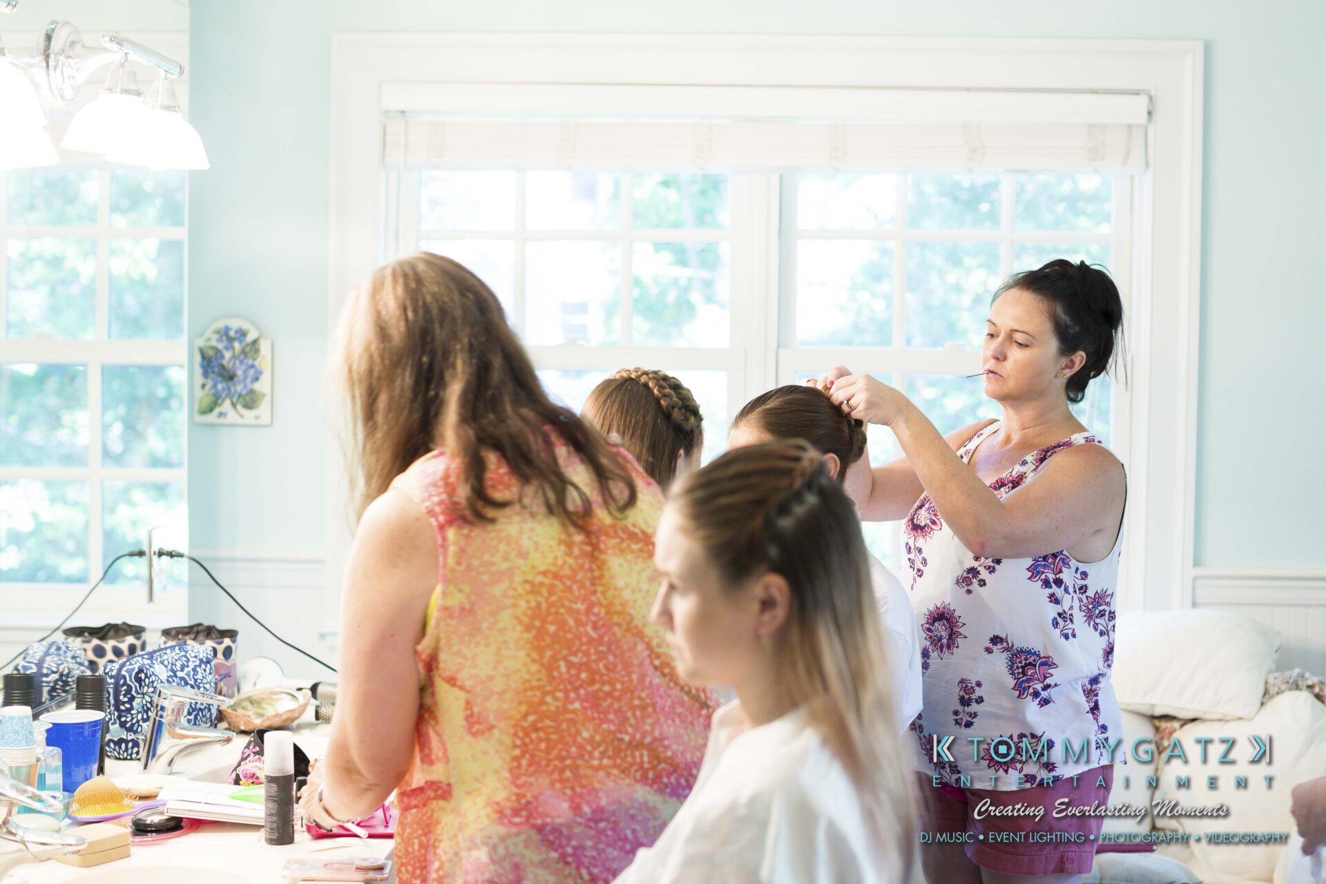 Ann-Marie-Alex-92317-PREP-Low-Res-WM-93 - TOMMY GATZ ENTERTAINMENT A woman is styling another woman's hair while two others sit nearby in a bright room with large windows. Various styling tools and products are on the table.