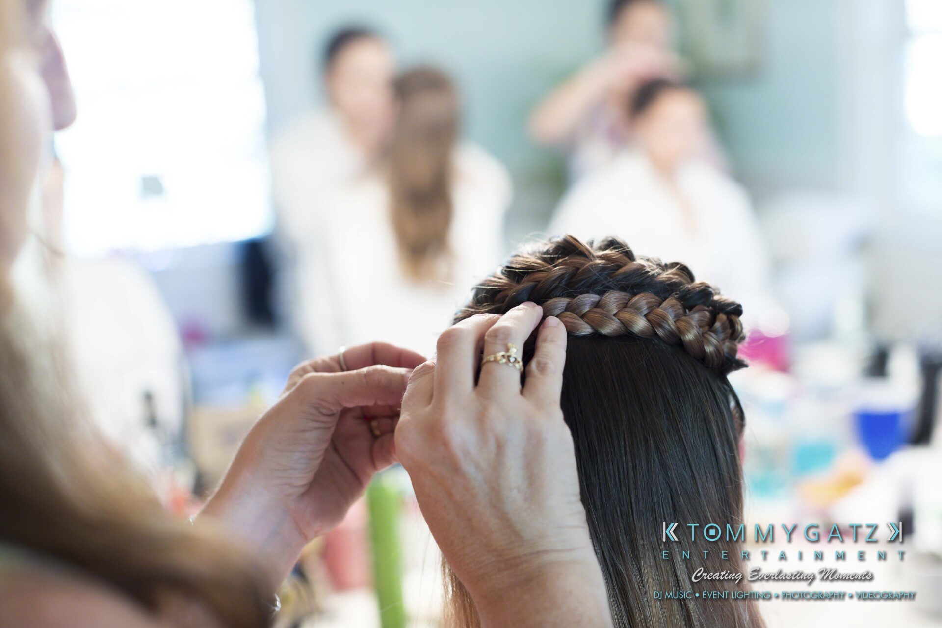 Ann-Marie-Alex-92317-PREP-Low-Res-WM-95 - TOMMY GATZ ENTERTAINMENT Hands styling a woman's hair into a braided updo, with blurred people and hair products visible in the background.