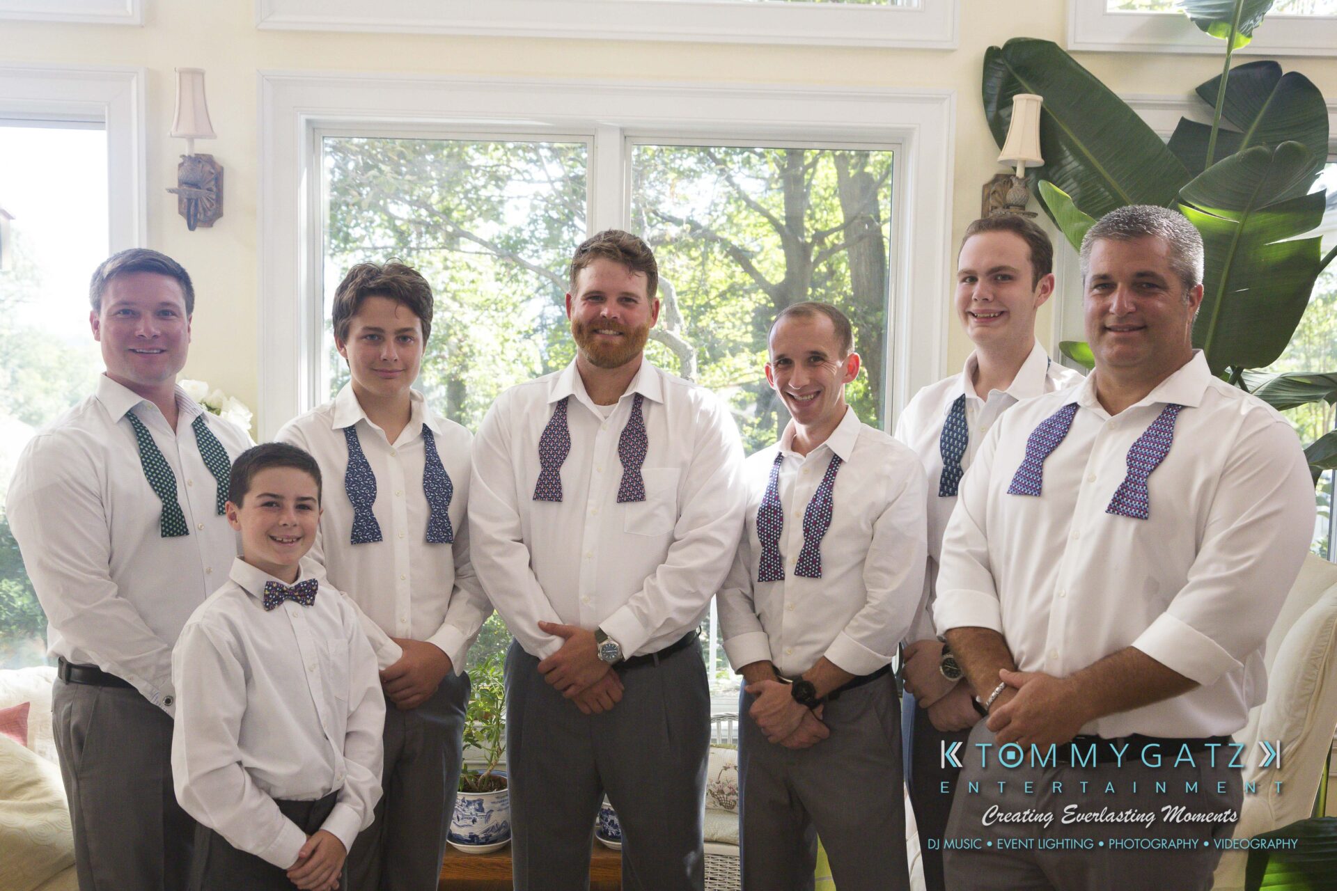 Ann-Marie-Alex-92317-PREP-Low-Res-WM-98 - TOMMY GATZ ENTERTAINMENT Seven men in dress shirts stand together, some wearing bow ties and some with untied ties, inside a room with large windows.