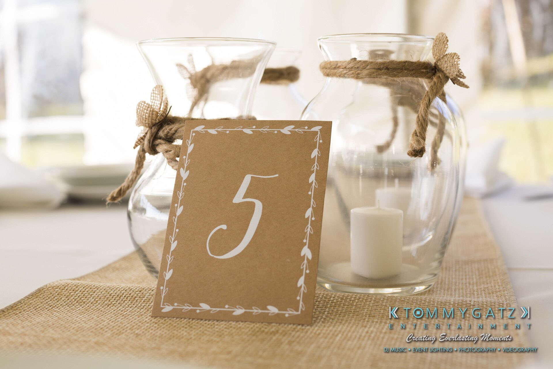 Ann-Marie-Alex-92317-RECEPTION-Low-Res-WM-1 - TOMMY GATZ ENTERTAINMENT Table decor featuring a card with the number 5, two glass vases tied with rope and filled with white candles, set on a burlap runner.
