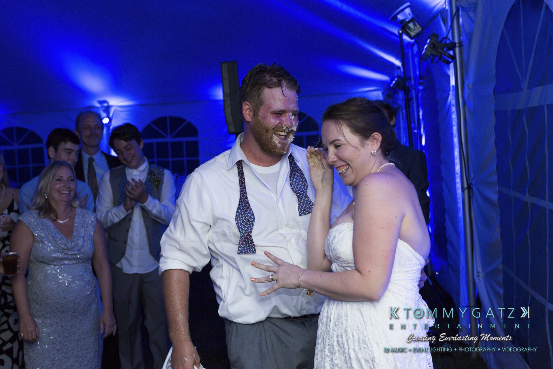 Ann-Marie-Alex-92317-RECEPTION-Low-Res-WM-100 - TOMMY GATZ ENTERTAINMENT A bride and groom laughing with cake on their faces at a wedding reception, surrounded by cheering guests in a blue-lit tent.