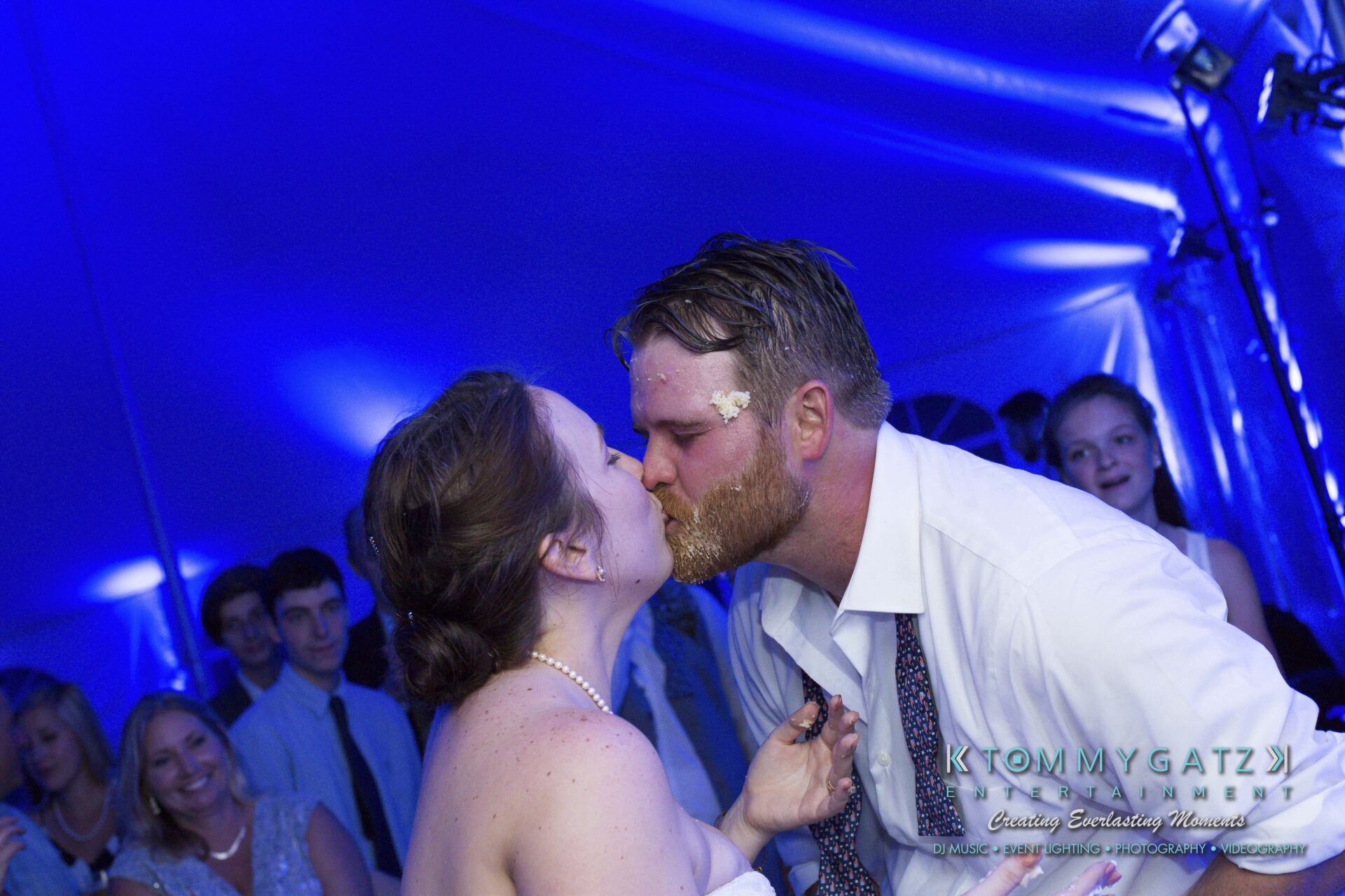 Ann-Marie-Alex-92317-RECEPTION-Low-Res-WM-103 - TOMMY GATZ ENTERTAINMENT A bride and groom share a kiss under blue lighting at a wedding reception, with cake on the groom's cheek. Guests watch in the background.