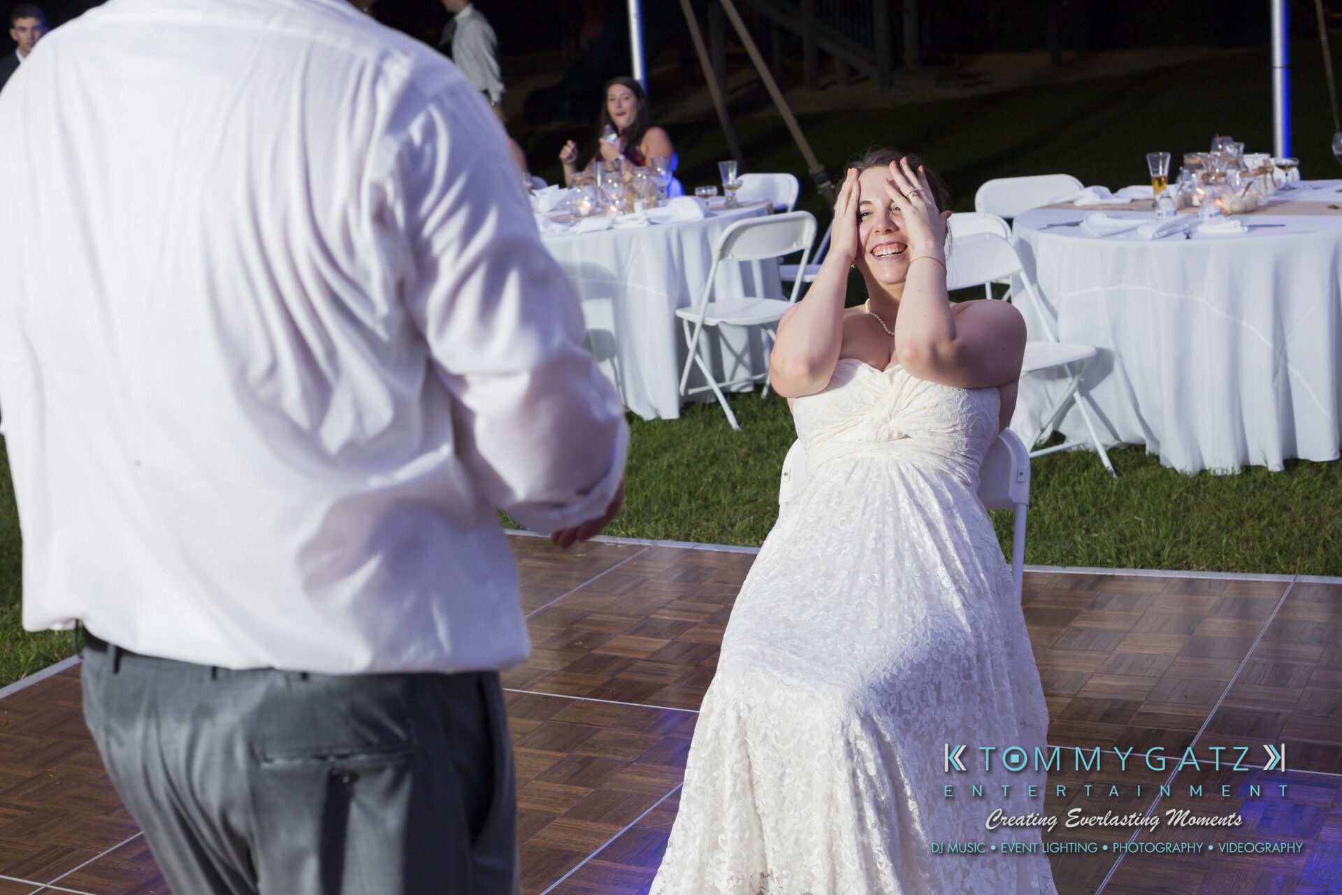 Ann-Marie-Alex-92317-RECEPTION-Low-Res-WM-105 - TOMMY GATZ ENTERTAINMENT A bride in a white dress sits on a chair on a dance floor, covering her face with her hands, as a man stands nearby. Tables with items are visible in the background.