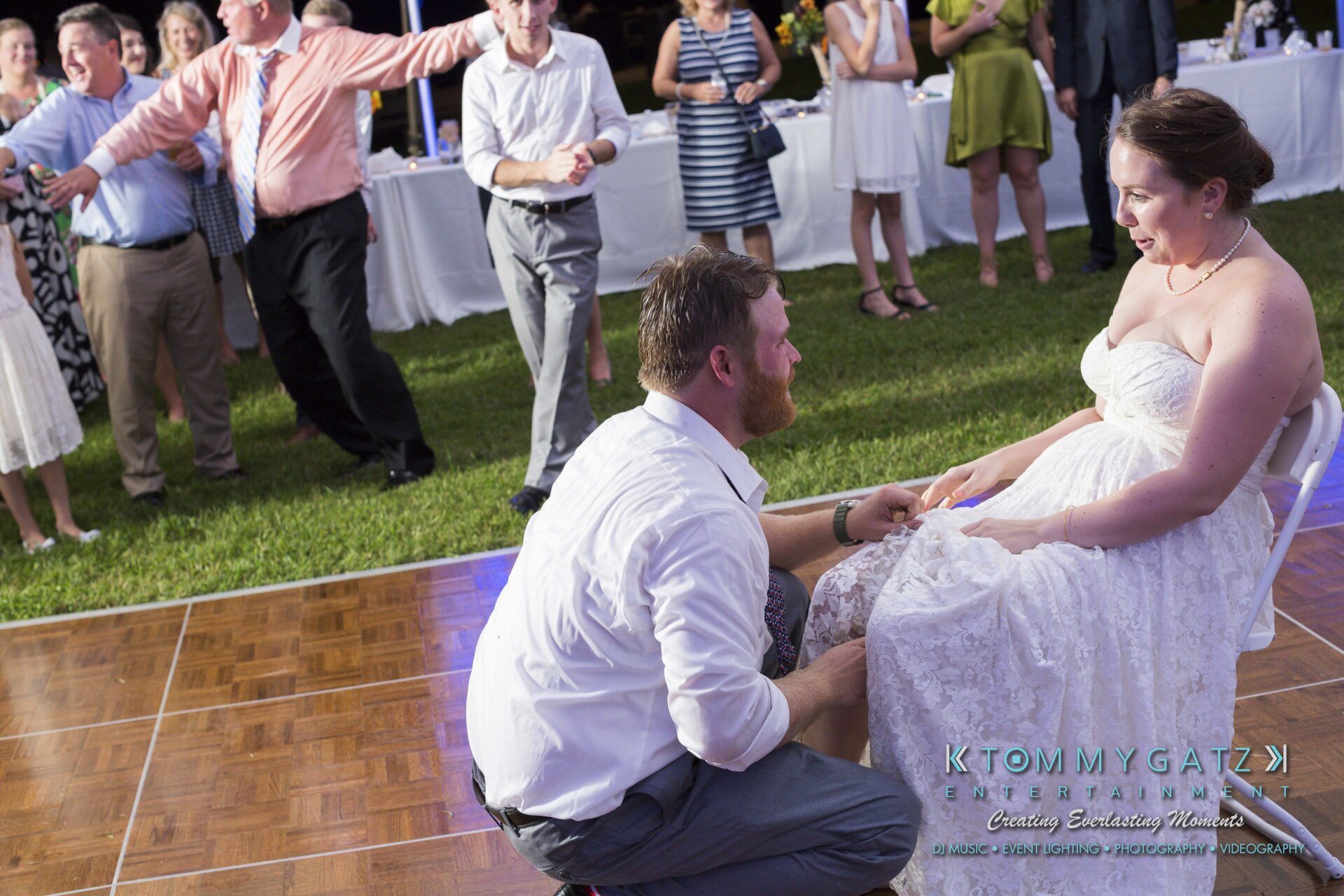Ann-Marie-Alex-92317-RECEPTION-Low-Res-WM-107 - TOMMY GATZ ENTERTAINMENT A man kneels in front of a seated woman on a dance floor, surrounded by people in formal attire at an outdoor event.