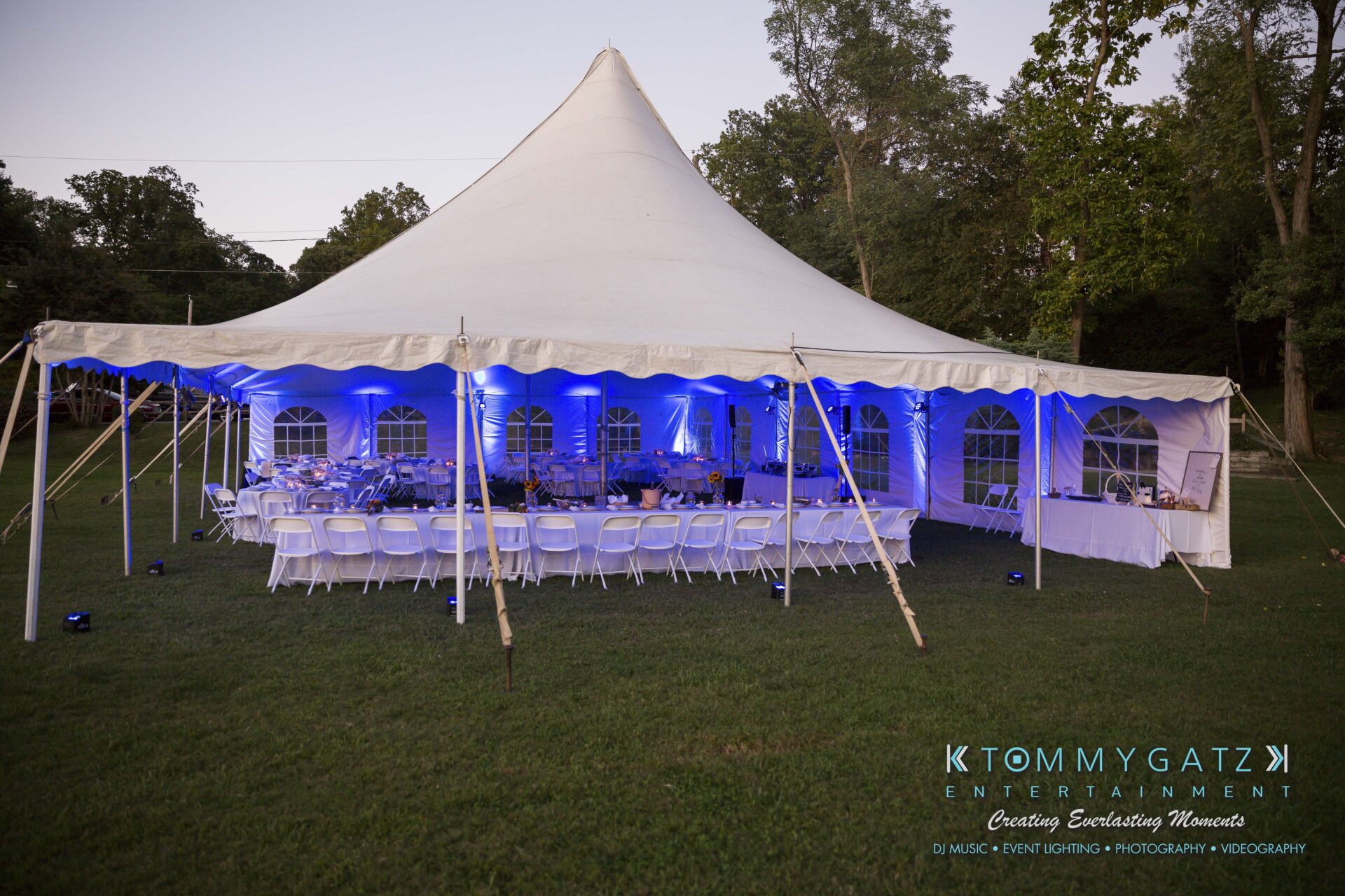 Ann-Marie-Alex-92317-RECEPTION-Low-Res-WM-11 - TOMMY GATZ ENTERTAINMENT A large white event tent with blue lighting, surrounded by trees, set up with white chairs and tables for an outdoor gathering.