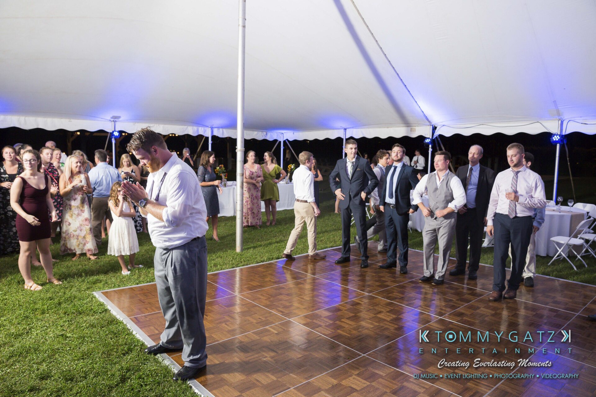 Ann-Marie-Alex-92317-RECEPTION-Low-Res-WM-110 - TOMMY GATZ ENTERTAINMENT A group of people at a wedding reception stand on a dance floor under a white tent. One man is giving a speech while guests watch attentively.