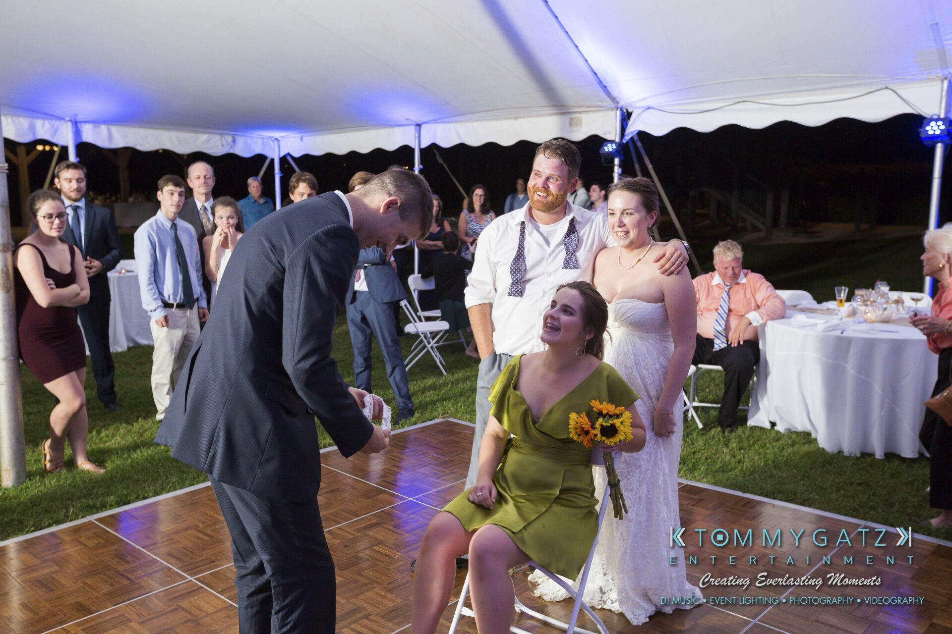 Ann-Marie-Alex-92317-RECEPTION-Low-Res-WM-114 - TOMMY GATZ ENTERTAINMENT A bride, groom, and guests watch a seated woman holding sunflowers, while a man in a suit stands nearby on a dance floor under a tent.