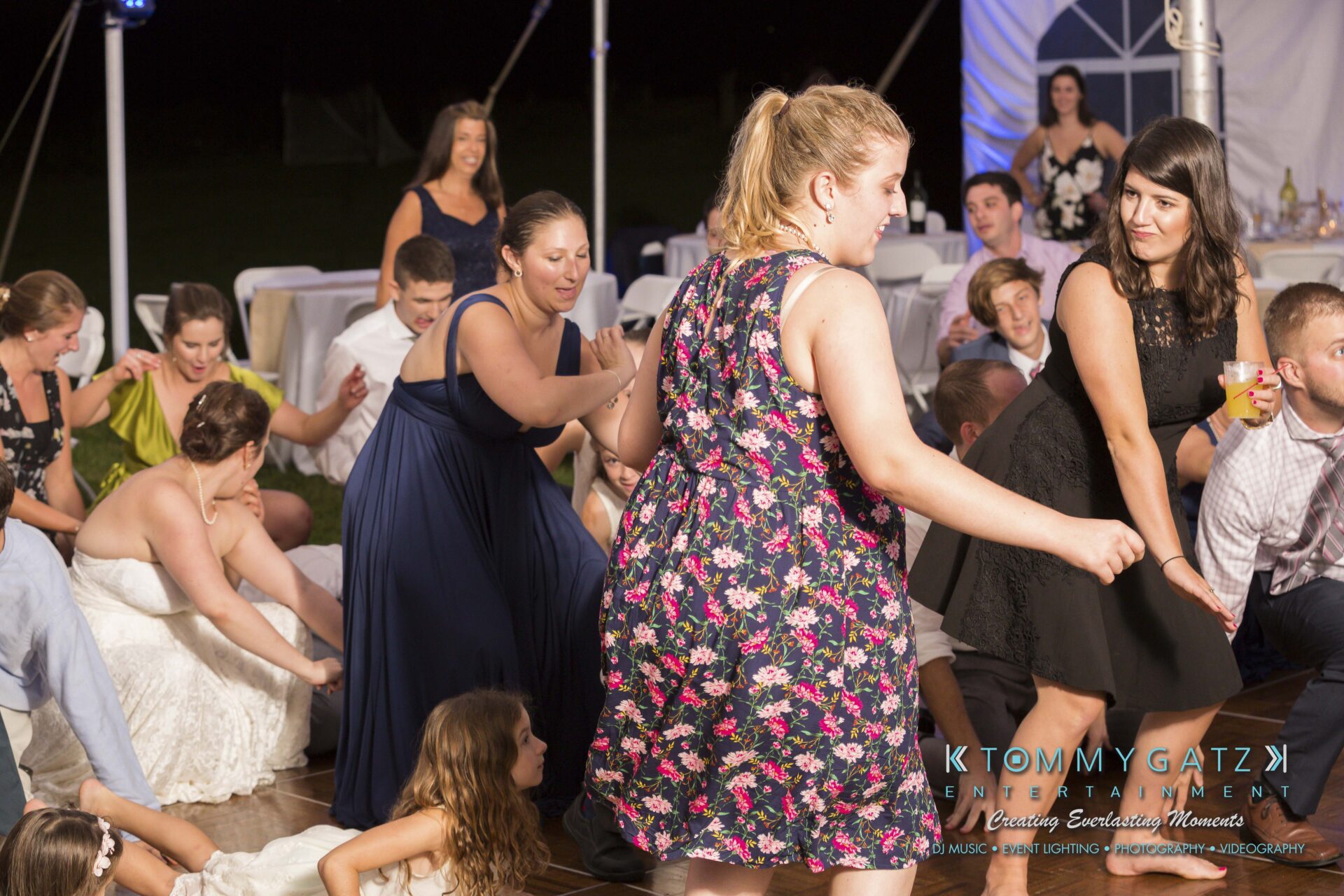 Ann-Marie-Alex-92317-RECEPTION-Low-Res-WM-122 - TOMMY GATZ ENTERTAINMENT A group of people dancing at an event on a wooden floor under a white tent. Some are sitting and clapping. The atmosphere is lively and festive.