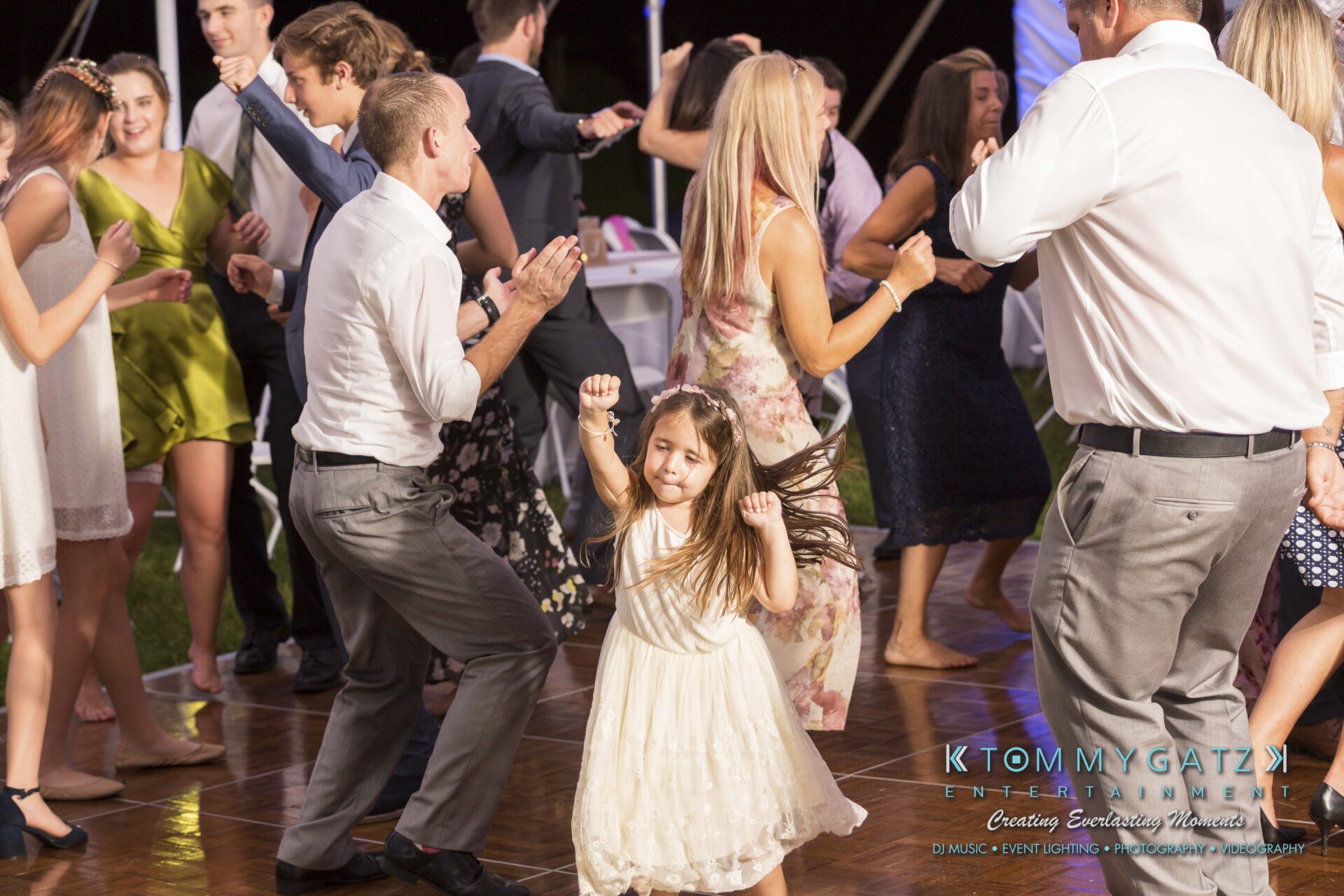 Ann-Marie-Alex-92317-RECEPTION-Low-Res-WM-123 - TOMMY GATZ ENTERTAINMENT A young girl in a white dress dances energetically on a wooden floor among a group of adults at a social event.