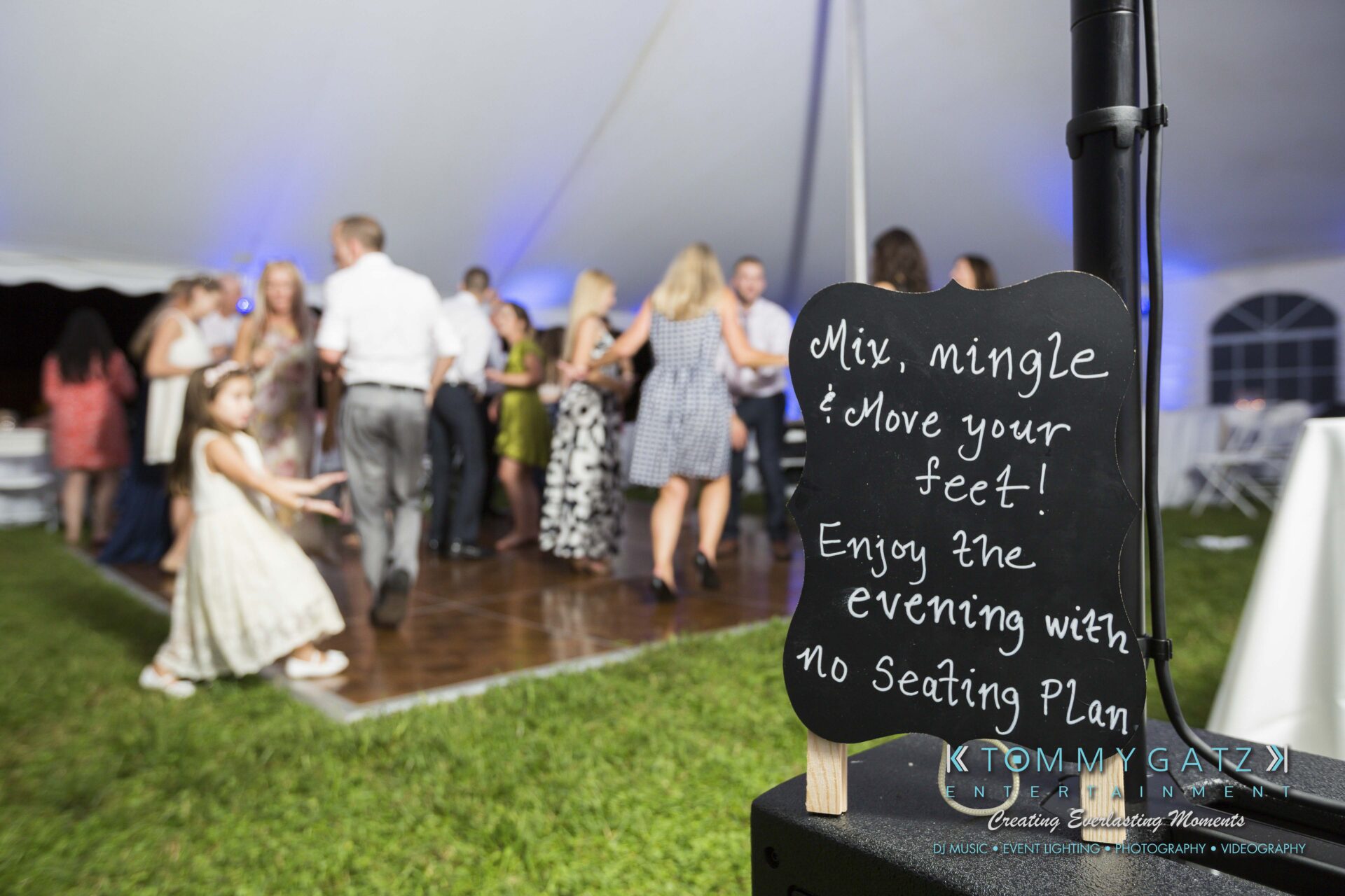 Ann-Marie-Alex-92317-RECEPTION-Low-Res-WM-125 - TOMMY GATZ ENTERTAINMENT Guests dancing on a wooden floor under a white tent, with a chalkboard sign that reads, "Mix, mingle & move your feet! Enjoy the evening with no seating plan.