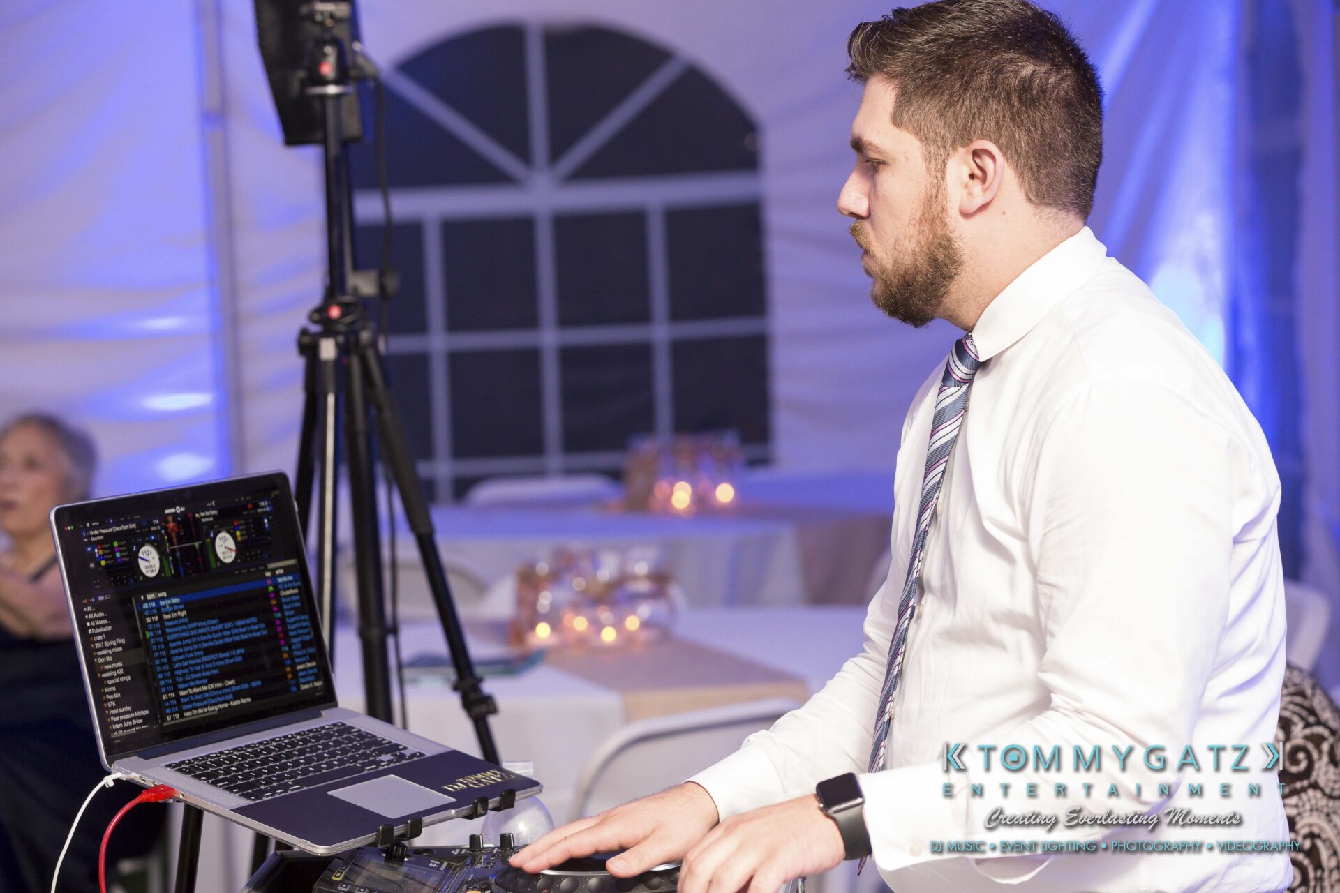 Ann-Marie-Alex-92317-RECEPTION-Low-Res-WM-126 - TOMMY GATZ ENTERTAINMENT A DJ in a white shirt and tie operates a laptop and DJ equipment at an event, with tables and guest in the background.
