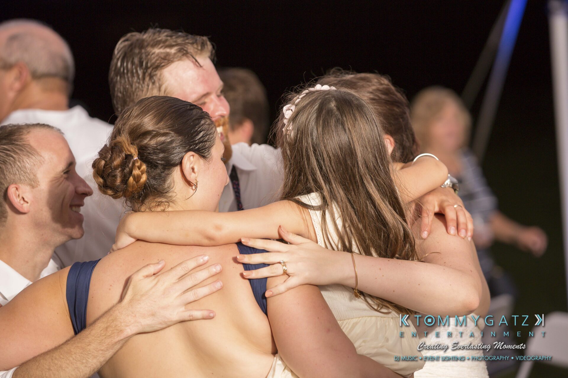 Ann-Marie-Alex-92317-RECEPTION-Low-Res-WM-129 - TOMMY GATZ ENTERTAINMENT A group hug involving several individuals, including a young girl with long hair, appears joyful. The setting is a nighttime event, and there's a logo in the corner.